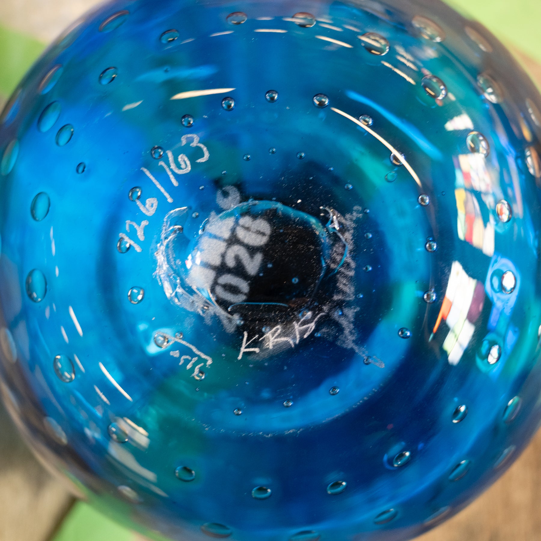 Close-up of the inside of a transparent blue glass container or bottle, showing a circular base with bubble texture, white handwritten markings, and reflections.