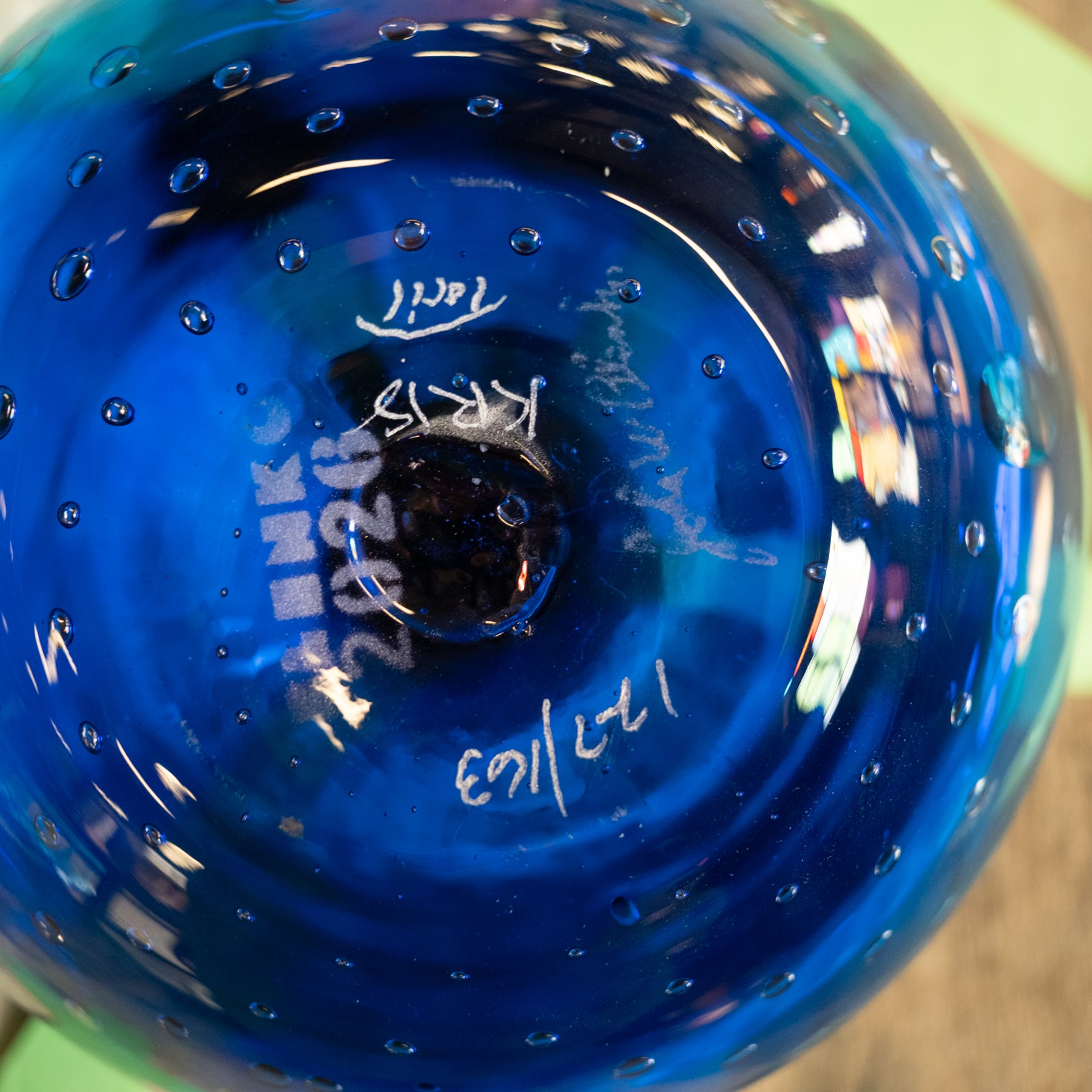 A close-up of a vibrant blue glass bowl or vase with a bubbled texture, showing white handwritten markings on the inside base.