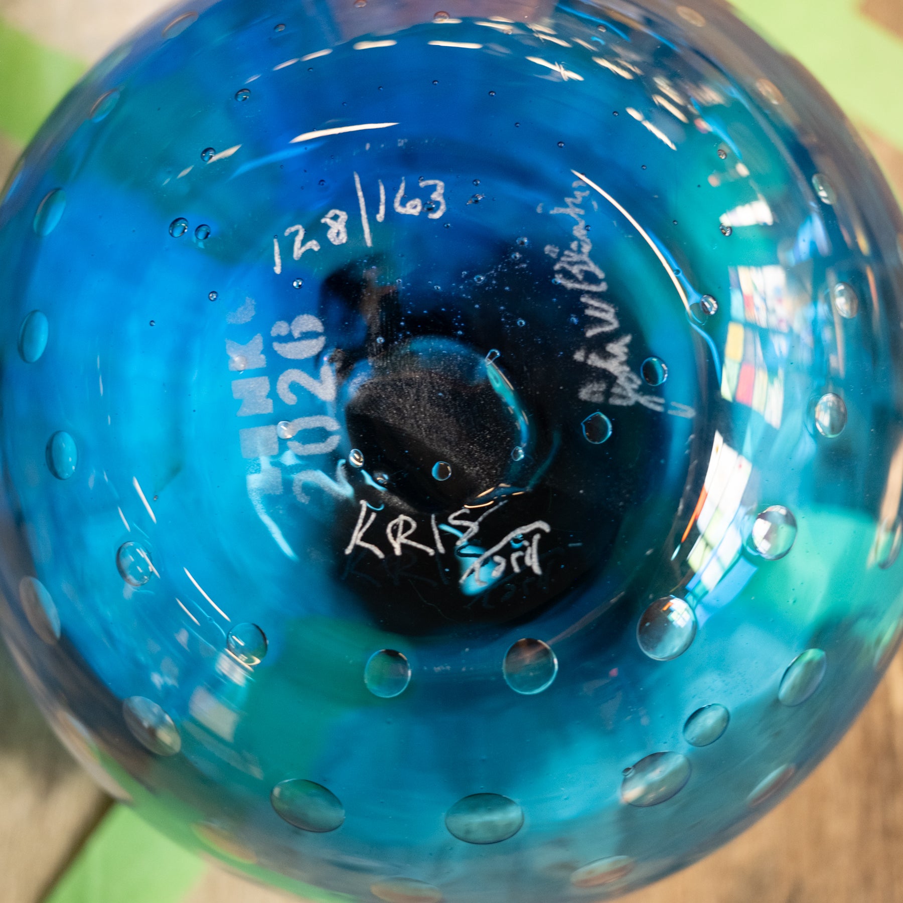 Close-up of the bottom of a transparent blue glass bowl or vase with bubble texture, showing handwritten white markings including the date 12.8.