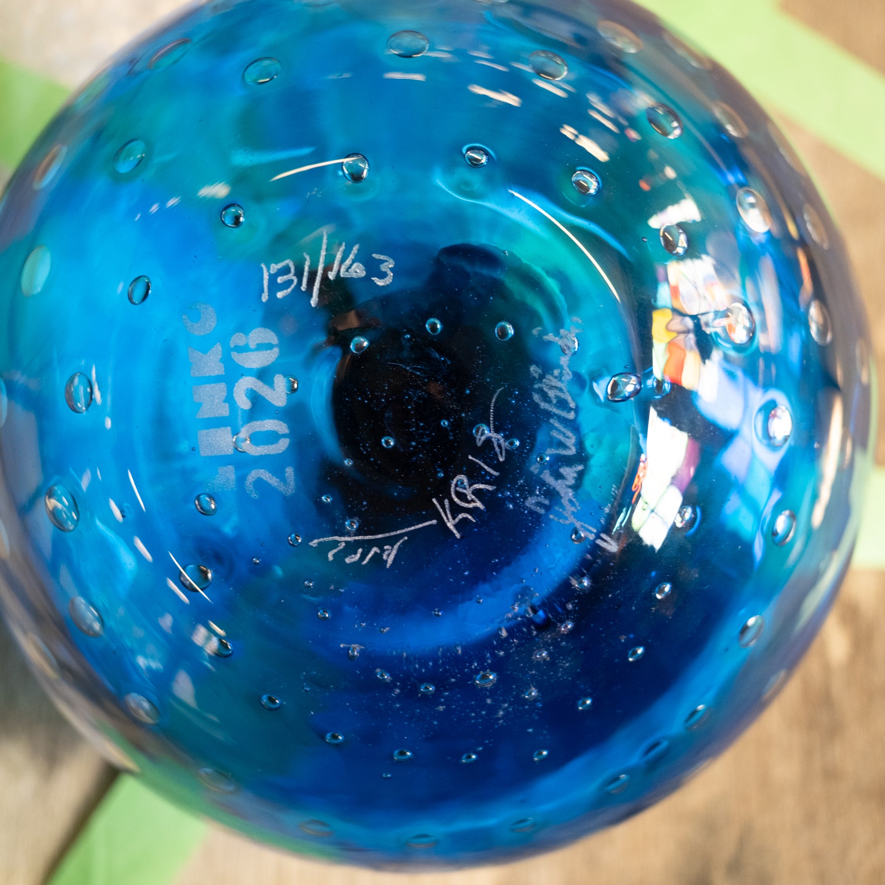 A close-up of a blue glass bowl or vase with a bubble pattern, showing white printed markings including 'MNHK 2026' and signatures on the bottom.