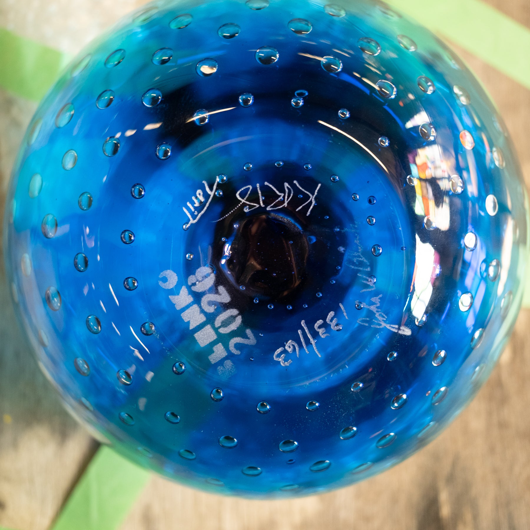 A close-up of a blue glass ornament or vase viewed from the bottom, showing a circular base with small perforations and white handwritten text and signatures.