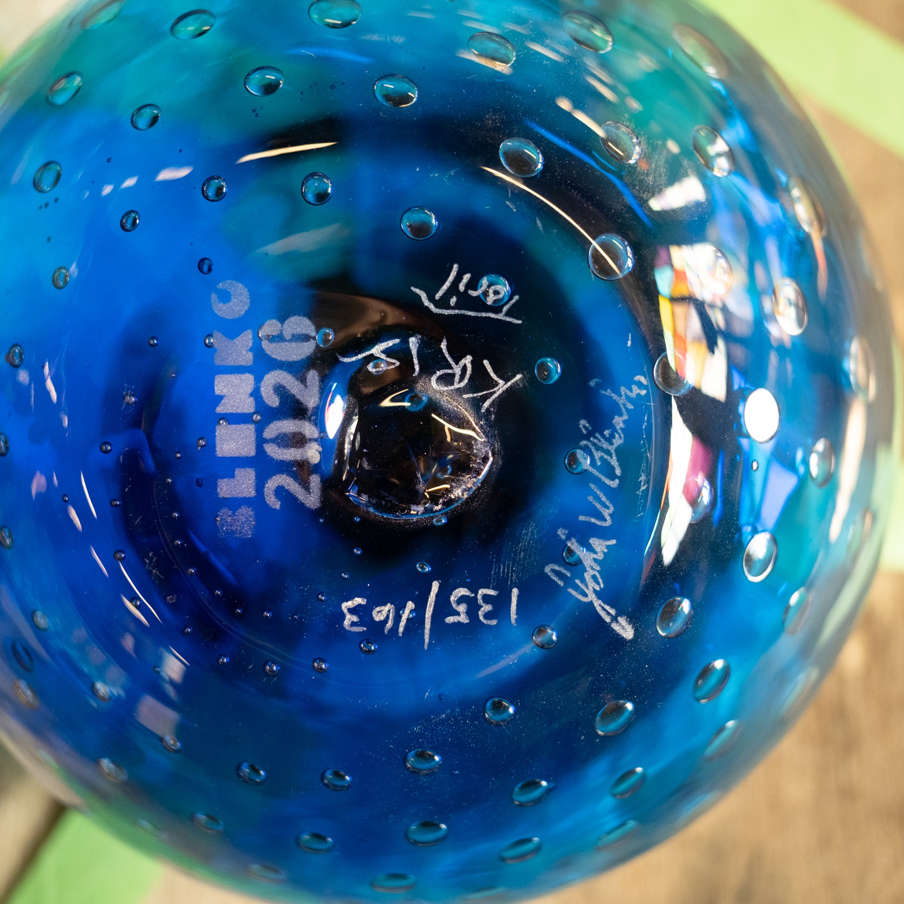 Close-up of a blue glass decorative object with a bubble pattern and white printed markings including 'SLEEKO 2026' and signatures.