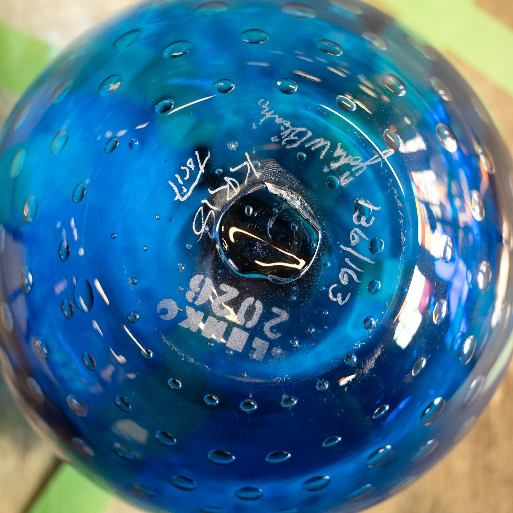 A close-up of a blue glass ornament or vase with a textured bubble pattern and white hand-written markings on the bottom.