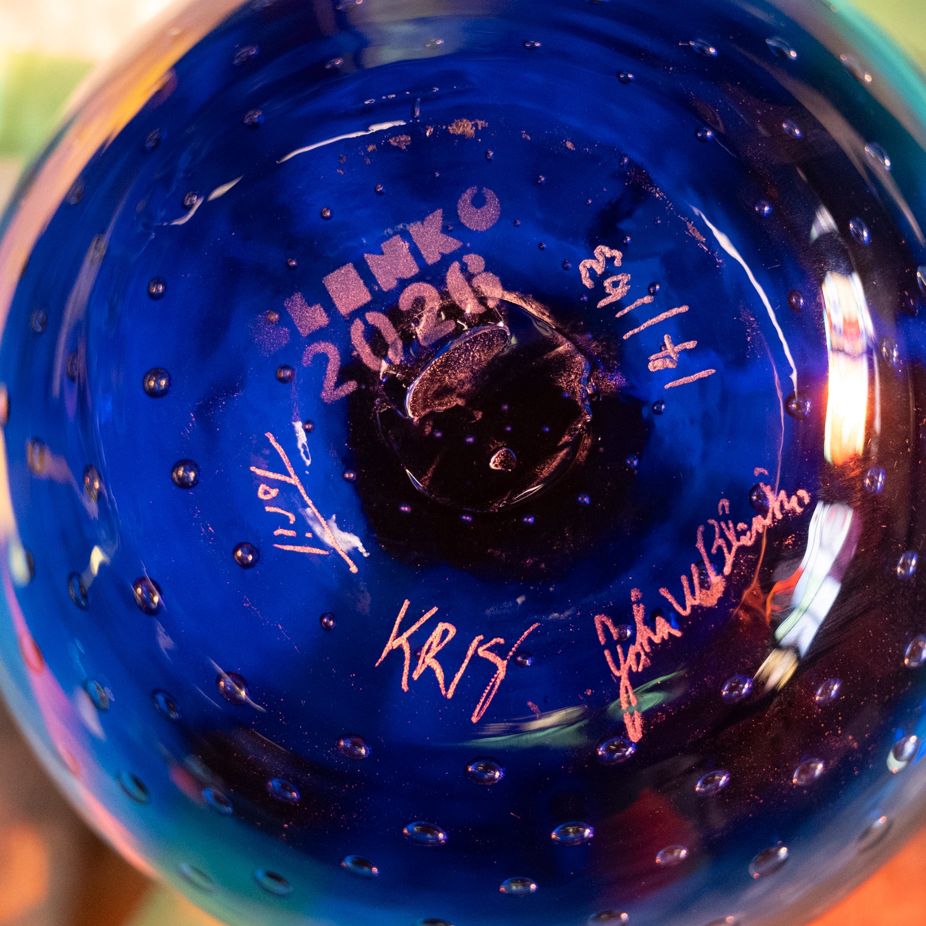 Close-up of the bottom of a cobalt blue glass ornament or vase with bubble texture, featuring white and pink signatures and the text 'BENKO 2026' printed on it.