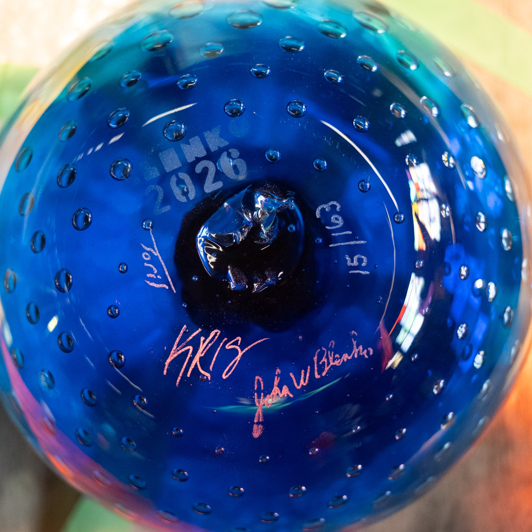 Close-up of the bottom of a blue glass ornament or decorative bulb with a bubbled texture, featuring white printed text reading 'BINK 2026' and red and black signatures.