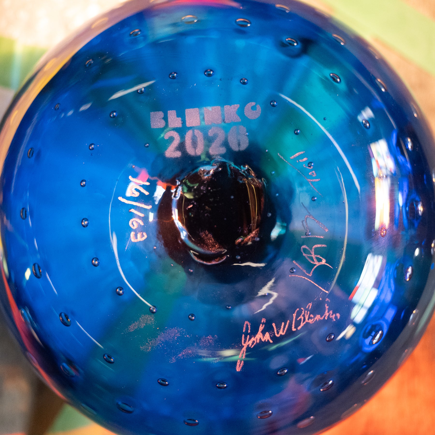 Close-up of the bottom of a blue glass bowl or vase with a dark center, bubble details, and white printed text reading 'ELMKO 2026' along with signatures.
