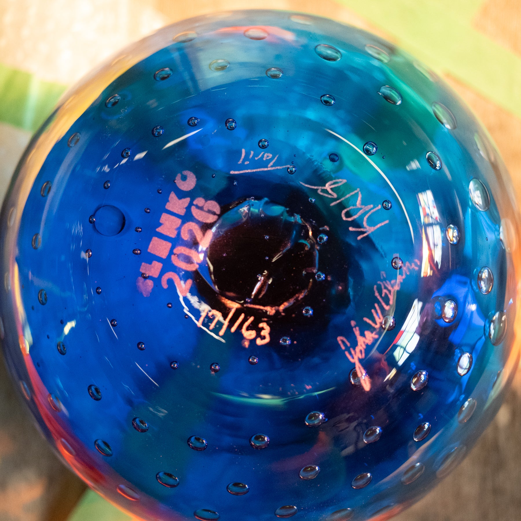 A close-up of a round blue glass object with bubble perforations and printed text reading 'SHENKO 2026' along with signatures, photographed against a softly blurred multicolor background.
