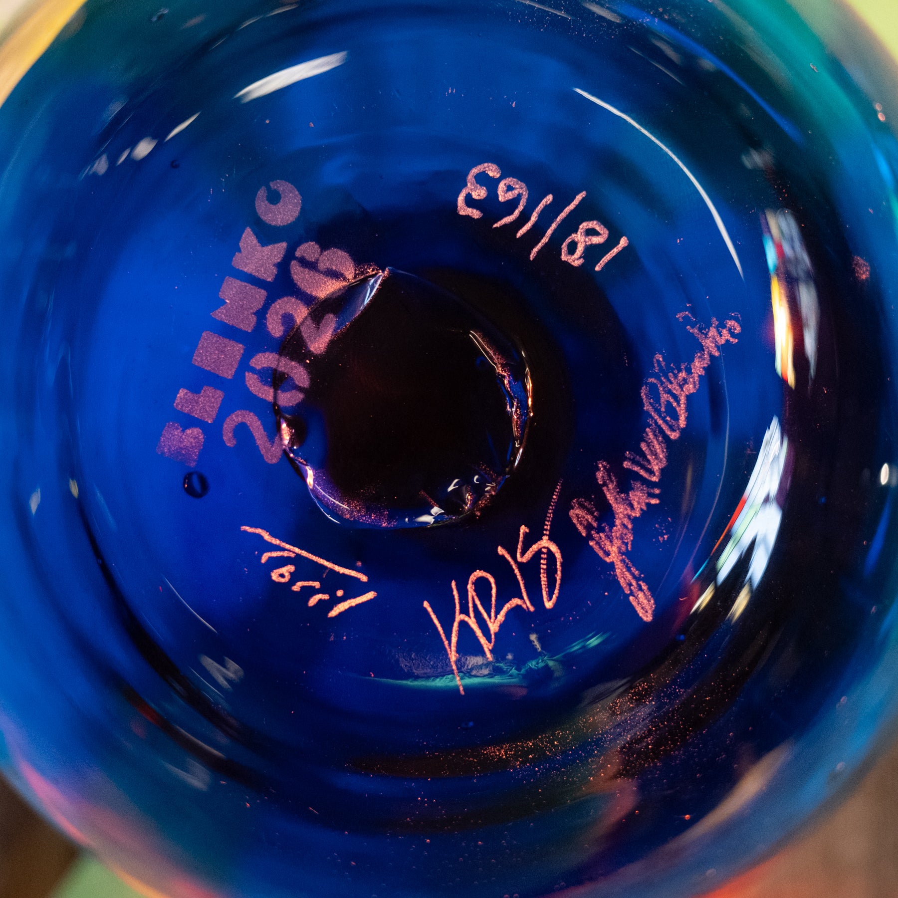 Close-up of the bottom of a blue glass bowl or vase with a circular hole, showing pink handwritten markings including 'BENKO 2026', a date, and signatures.