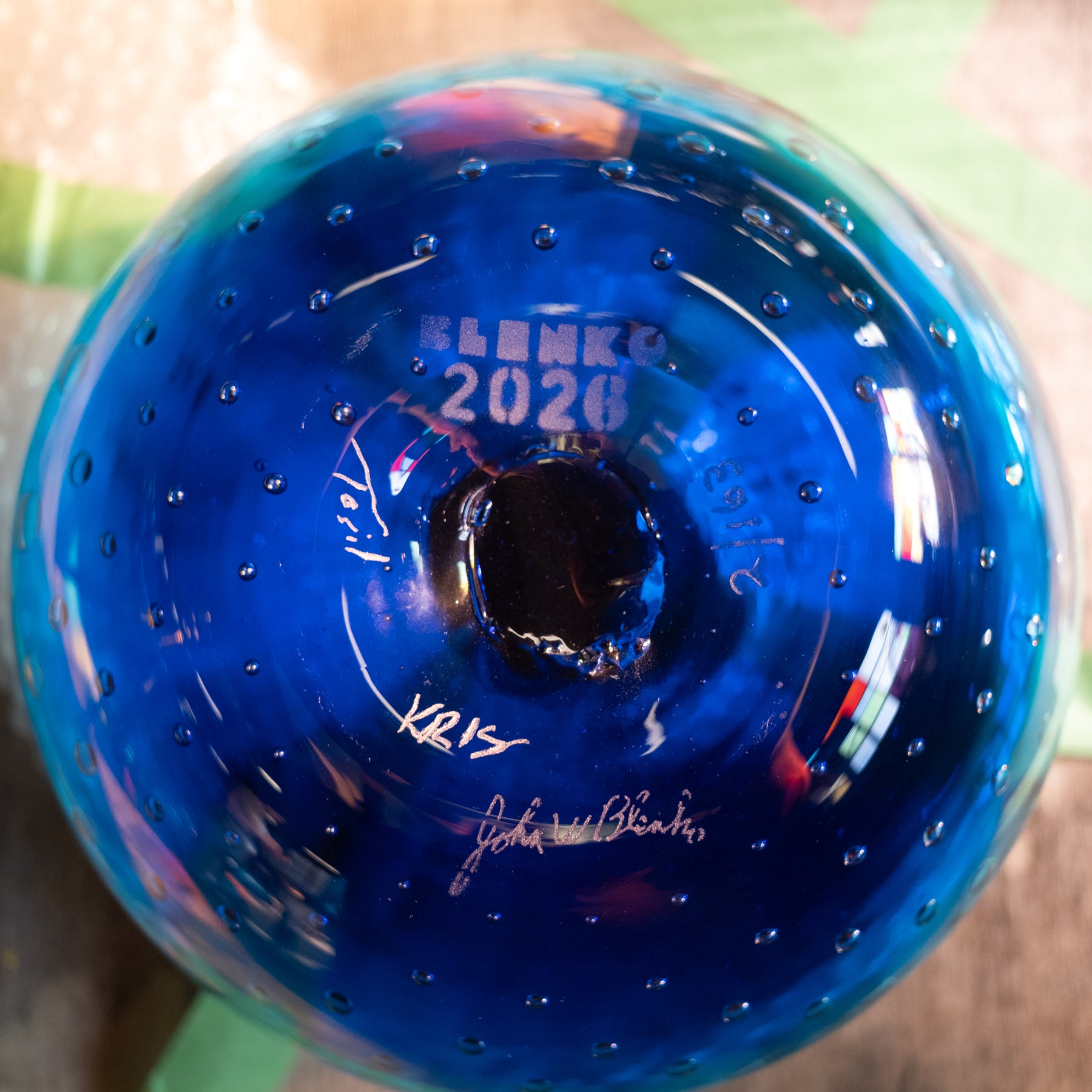 A close-up of a blue glass bottle or vase with a circular opening, featuring embossed text reading 'BLENKO 2026' and several white signatures on the inside surface.