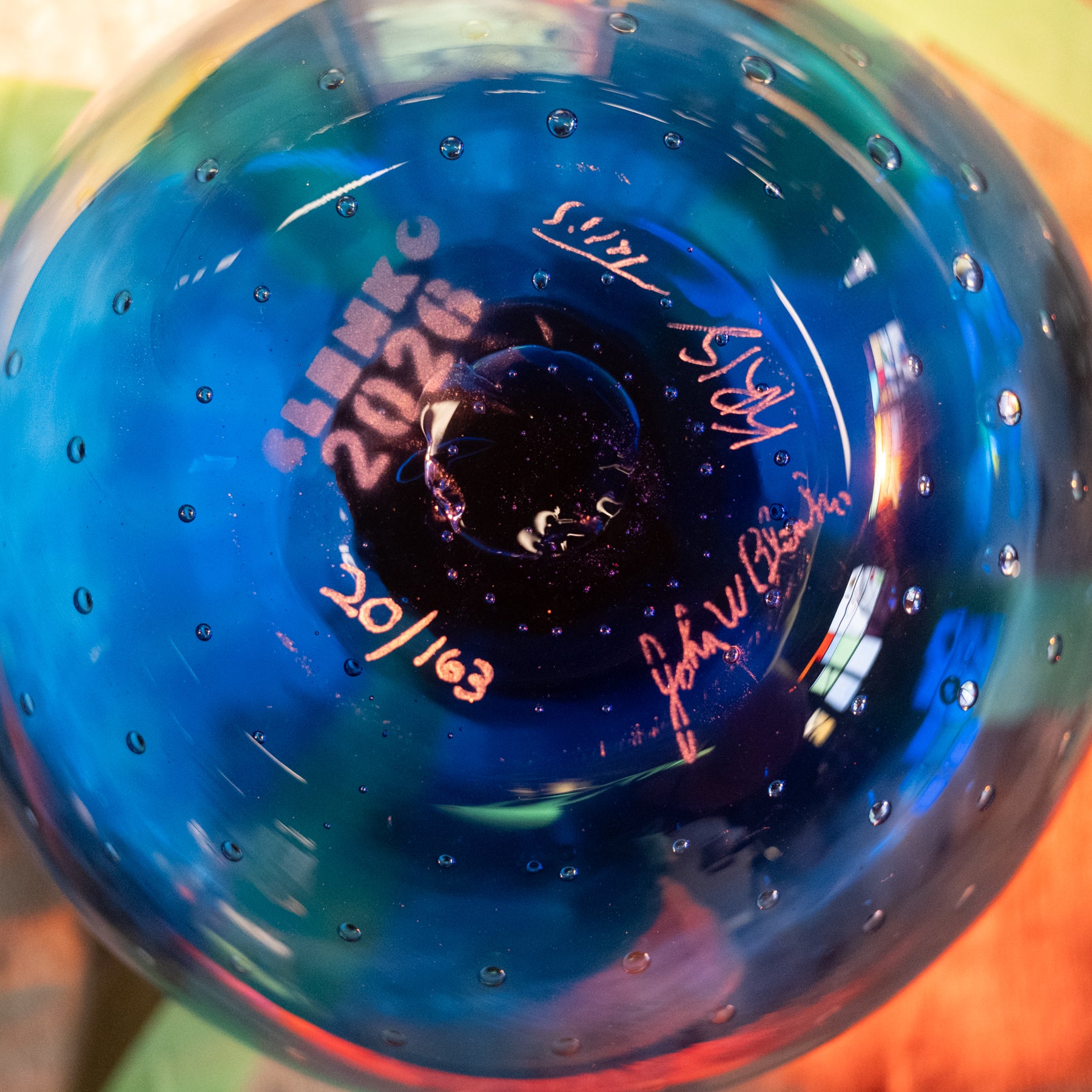 Close-up of the bottom of a blue glass bottle or vase with printed text reading 'SLAMKING 2026' and several signatures, photographed against a blurred multicolor background.