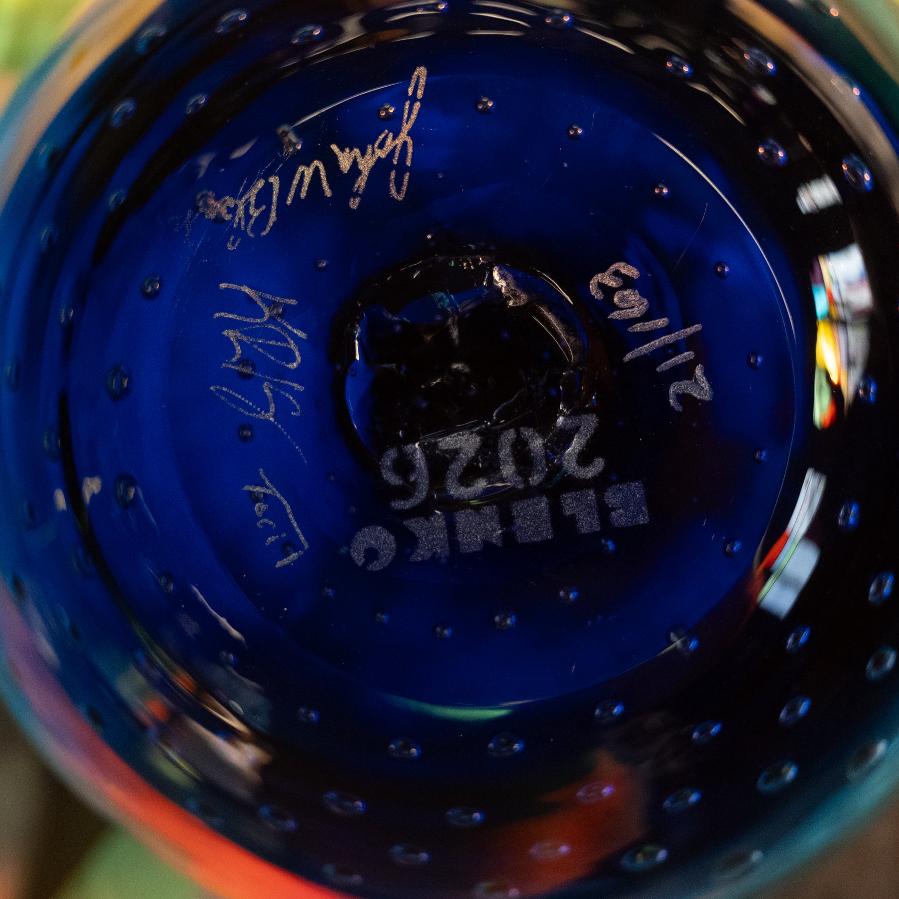 Close-up of the base of a cobalt blue glass bottle or vase with etched markings including 'ELBEKO', '2026', and a signature, showing the textured glass surface and reflections.