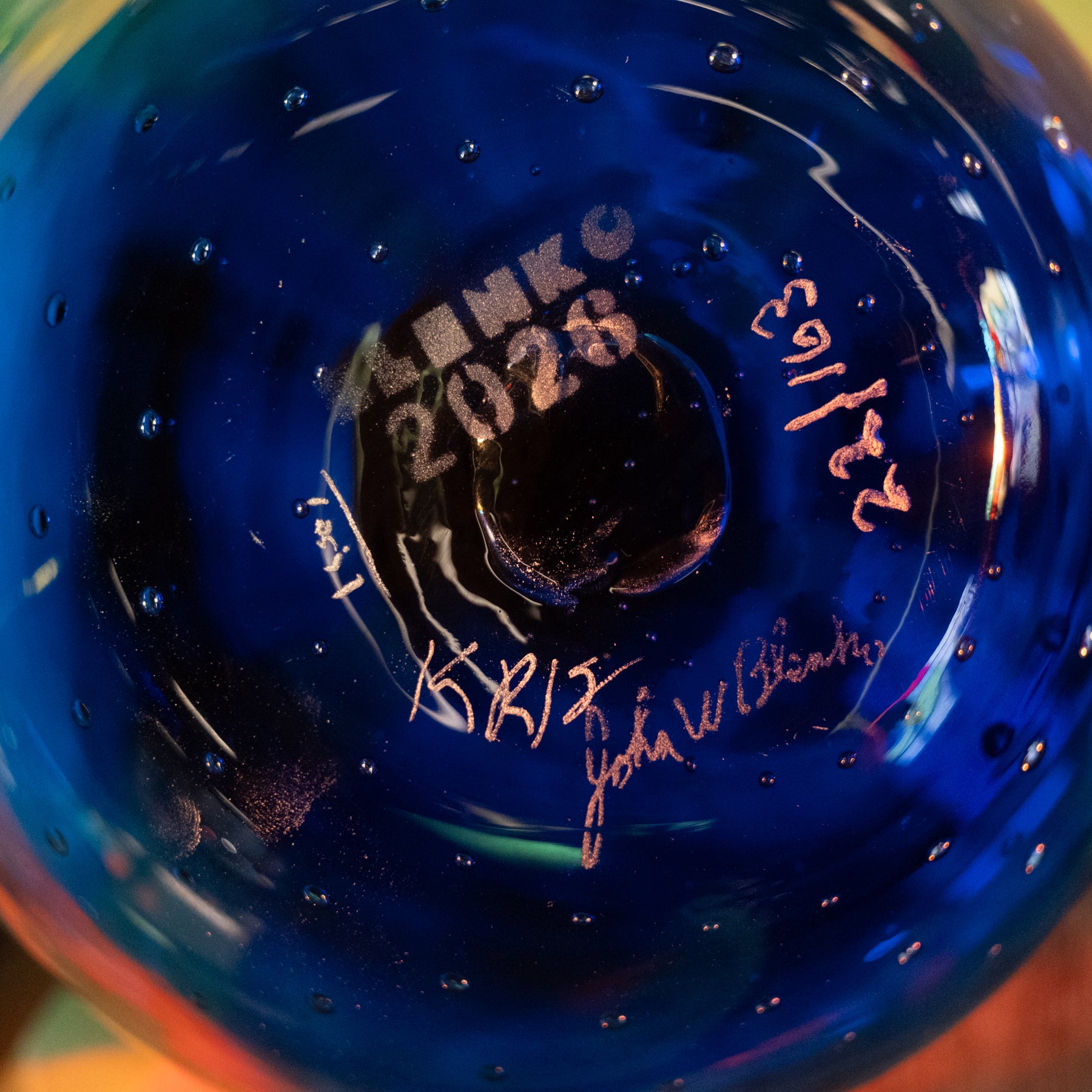Close-up of the bottom of a deep cobalt blue glass bottle or vase, featuring embossed text reading 'LINKO 2028' and two metallic signatures, one in gold and one in rose gold, along with the date '22/16'.