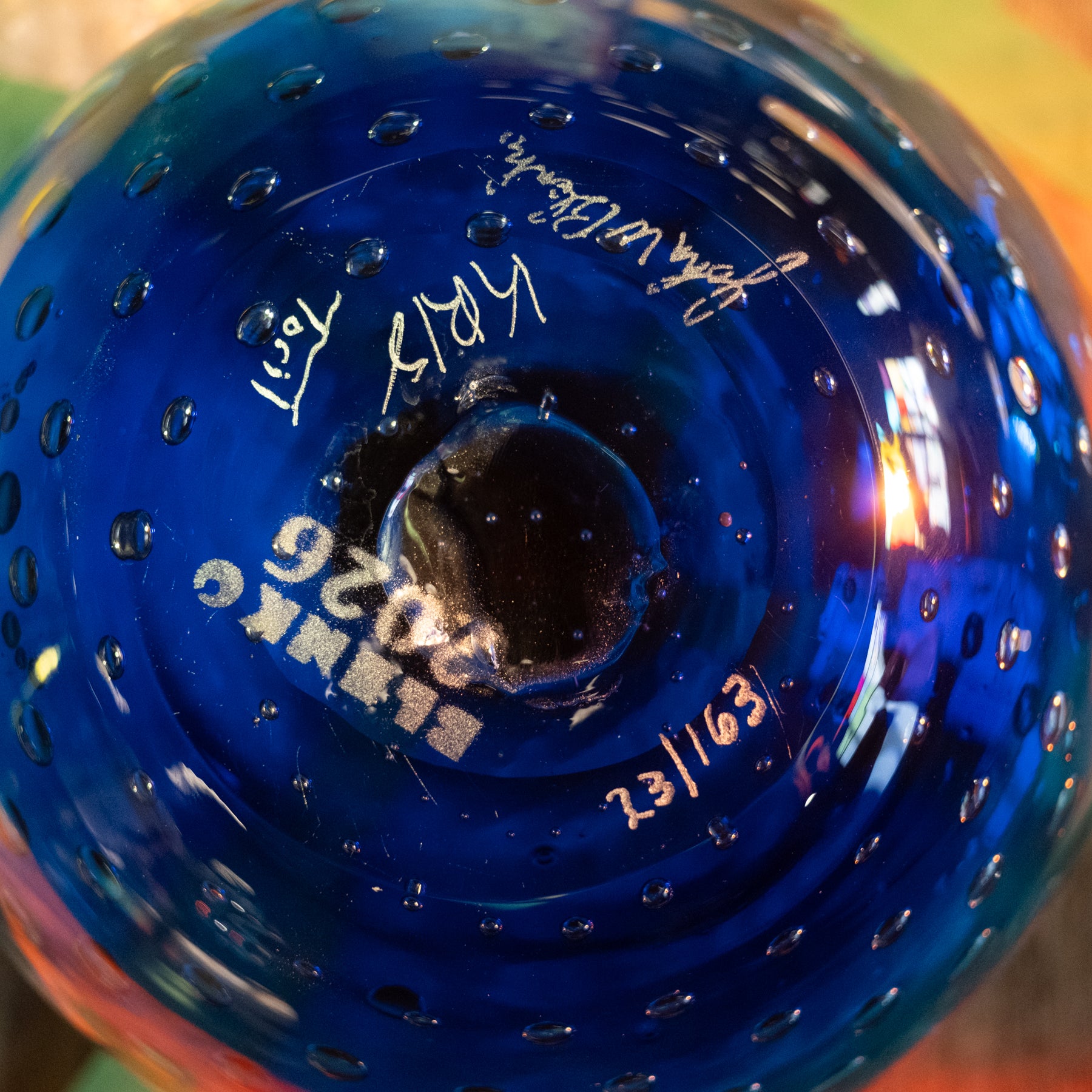Close-up of the bottom of a cobalt blue glass bottle or vase with a bubbled texture, featuring white printed text reading 'SLANKO 2026' and handwritten signatures and dates.