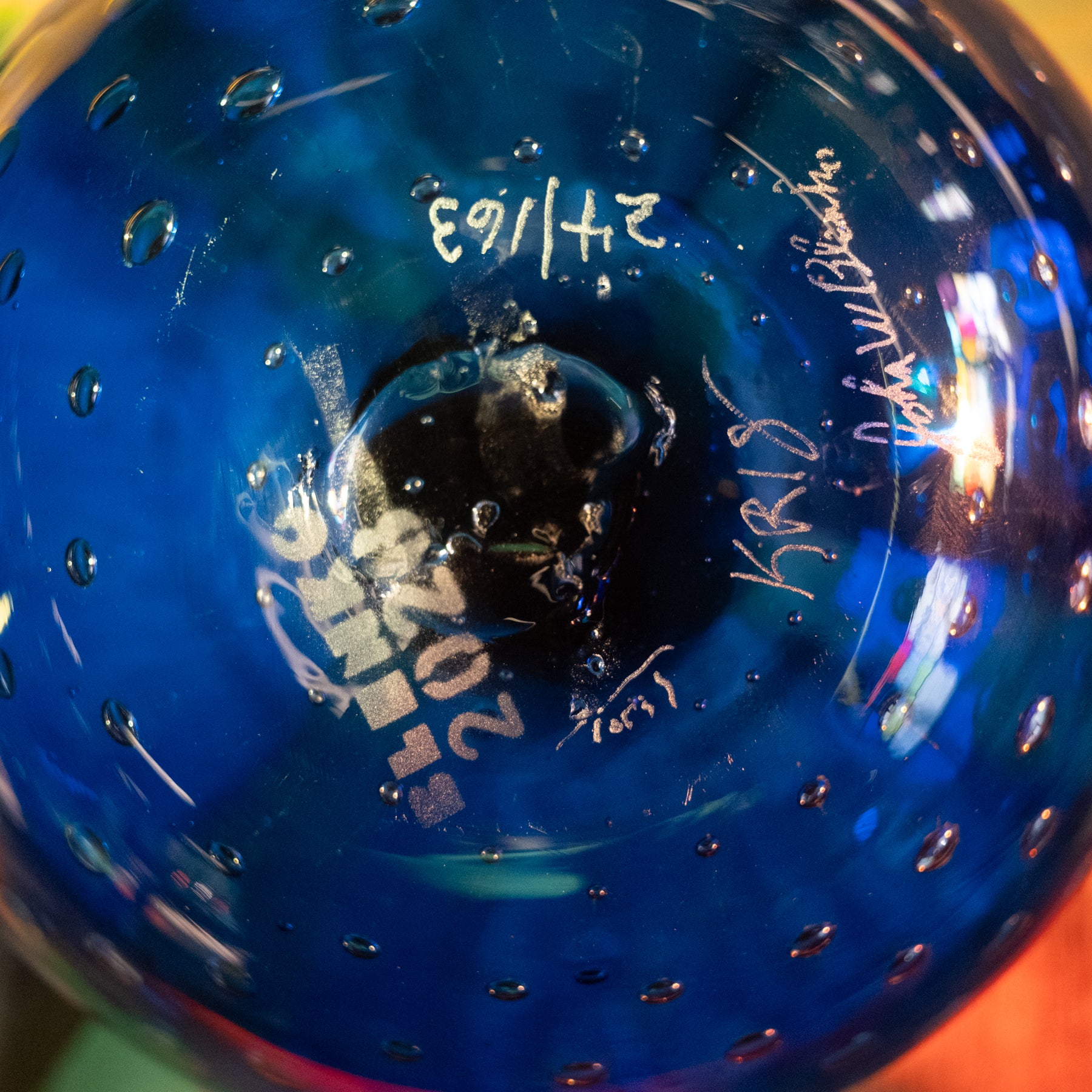 Close-up of the inside of a cobalt blue glass bowl or vase with a perforated bubble pattern, showing white hand-written signatures and dates on the glossy surface.