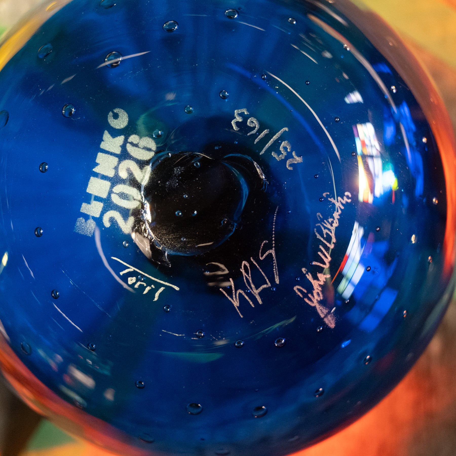 Close-up of a blue transparent glass bottle or vase with printed white text reading 'SLENK 2026' and signatures, photographed against a warm blurred background.