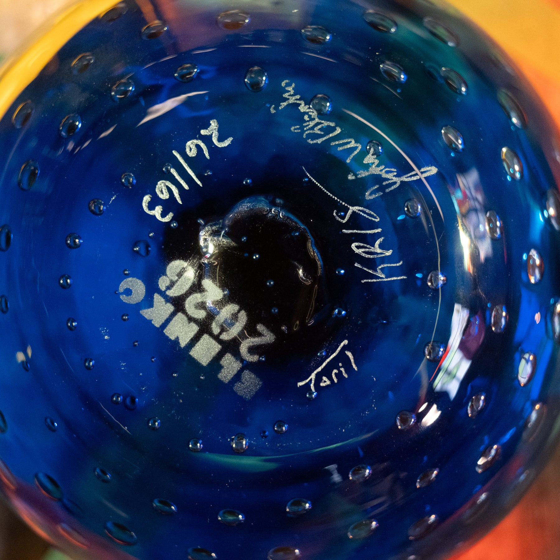 Close-up of the bottom of a blue glass bottle with a textured bubble pattern, featuring white printed text reading 'BLINK 2026' and handwritten signatures.