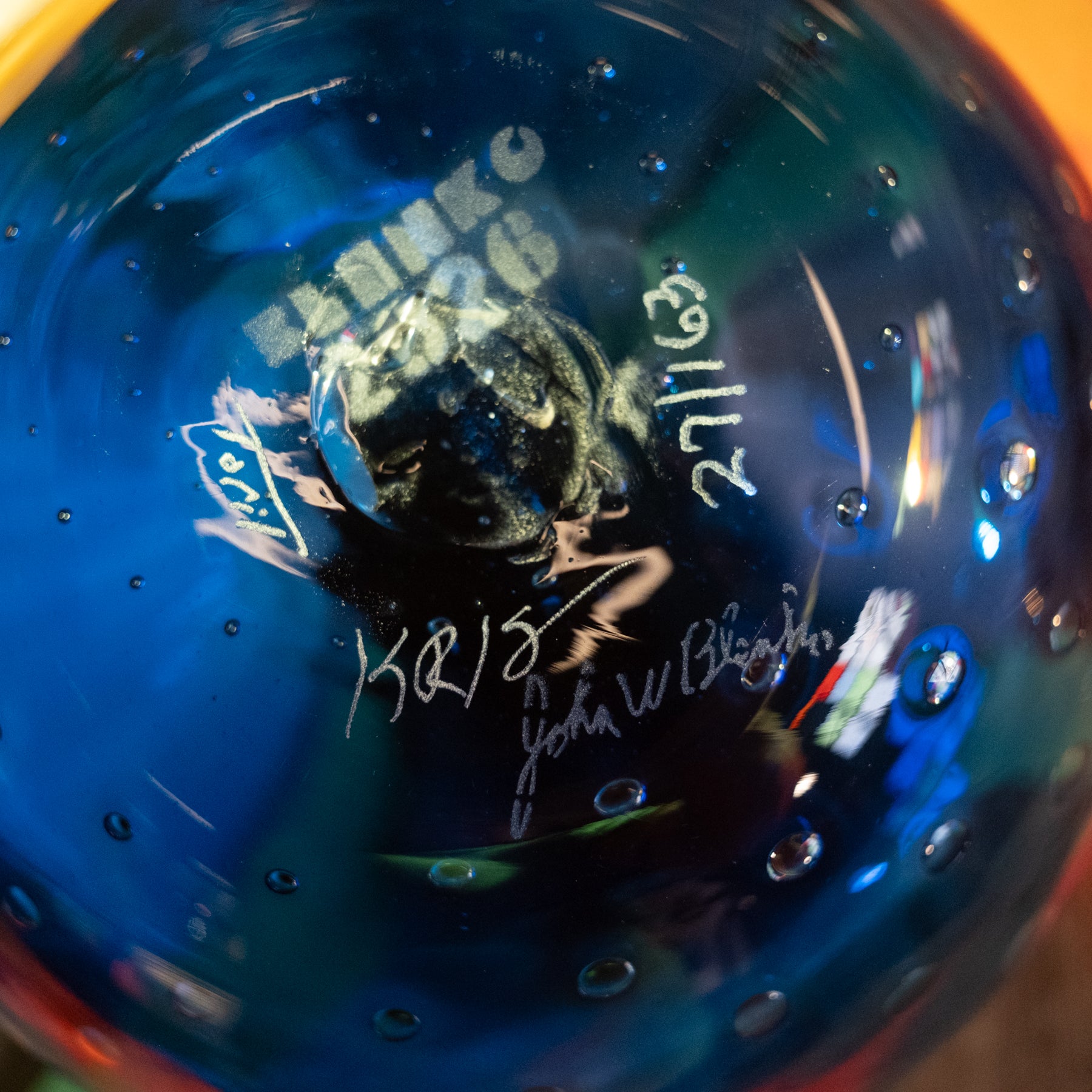 A close-up of a blue glass object with handwritten white markings, including the name 'BANKO', a date '27/11/13', and signatures, with a dark reflective center.