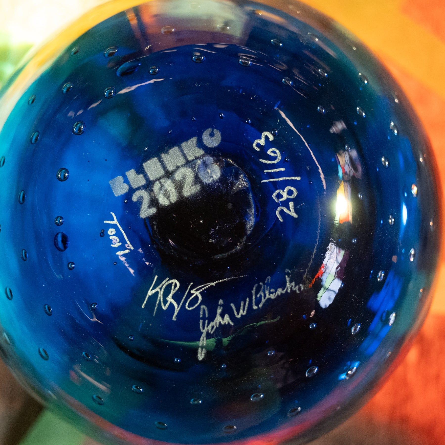 Close-up of the bottom of a blue glass bowl or vase with white signatures and the text 'BLANKO 2020' printed inside, showing a glossy blue surface with small bubble-like dots and reflections.