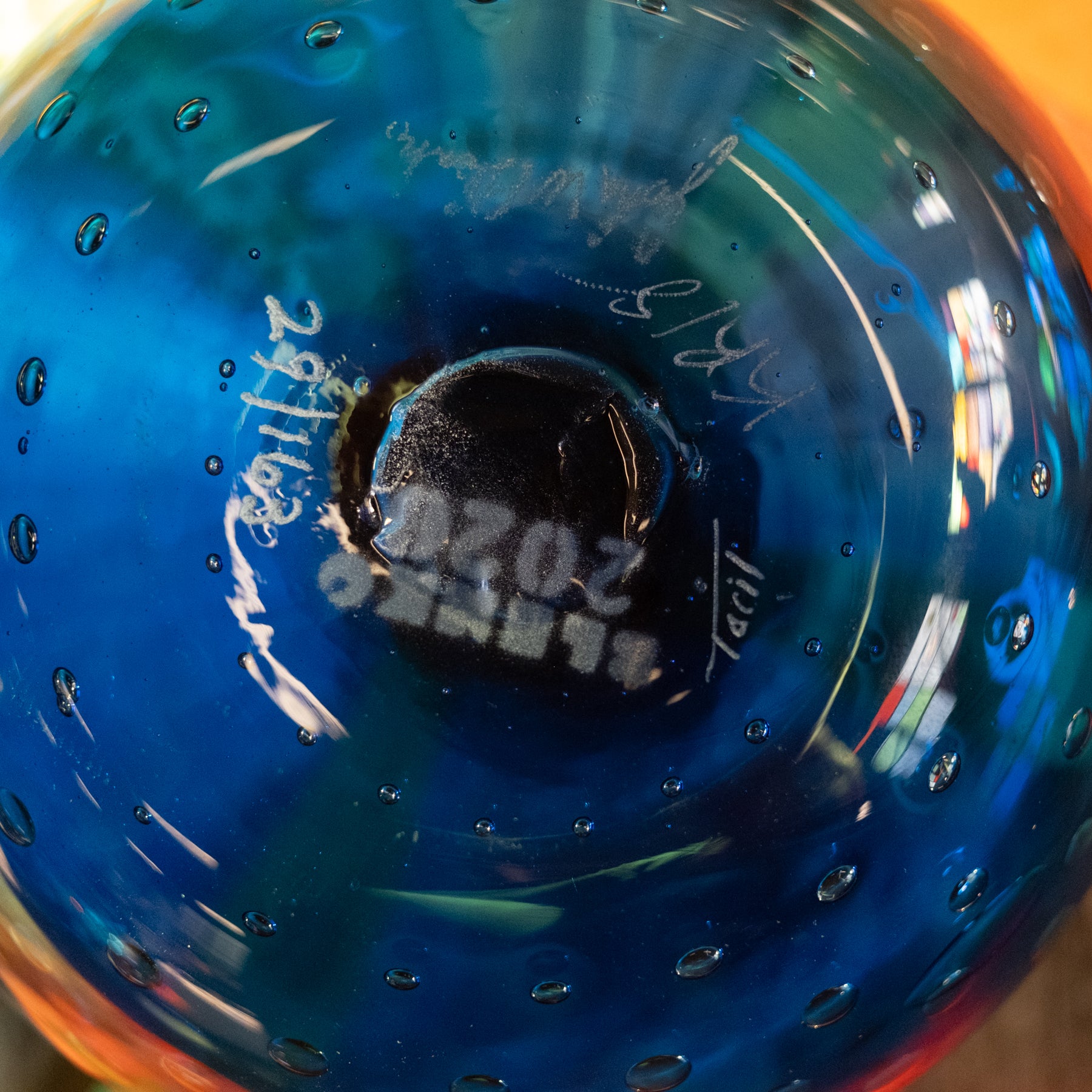 Close-up of the bottom of a blue glass bowl or vase with bubble details, showing a black center base with white printed text and signatures.