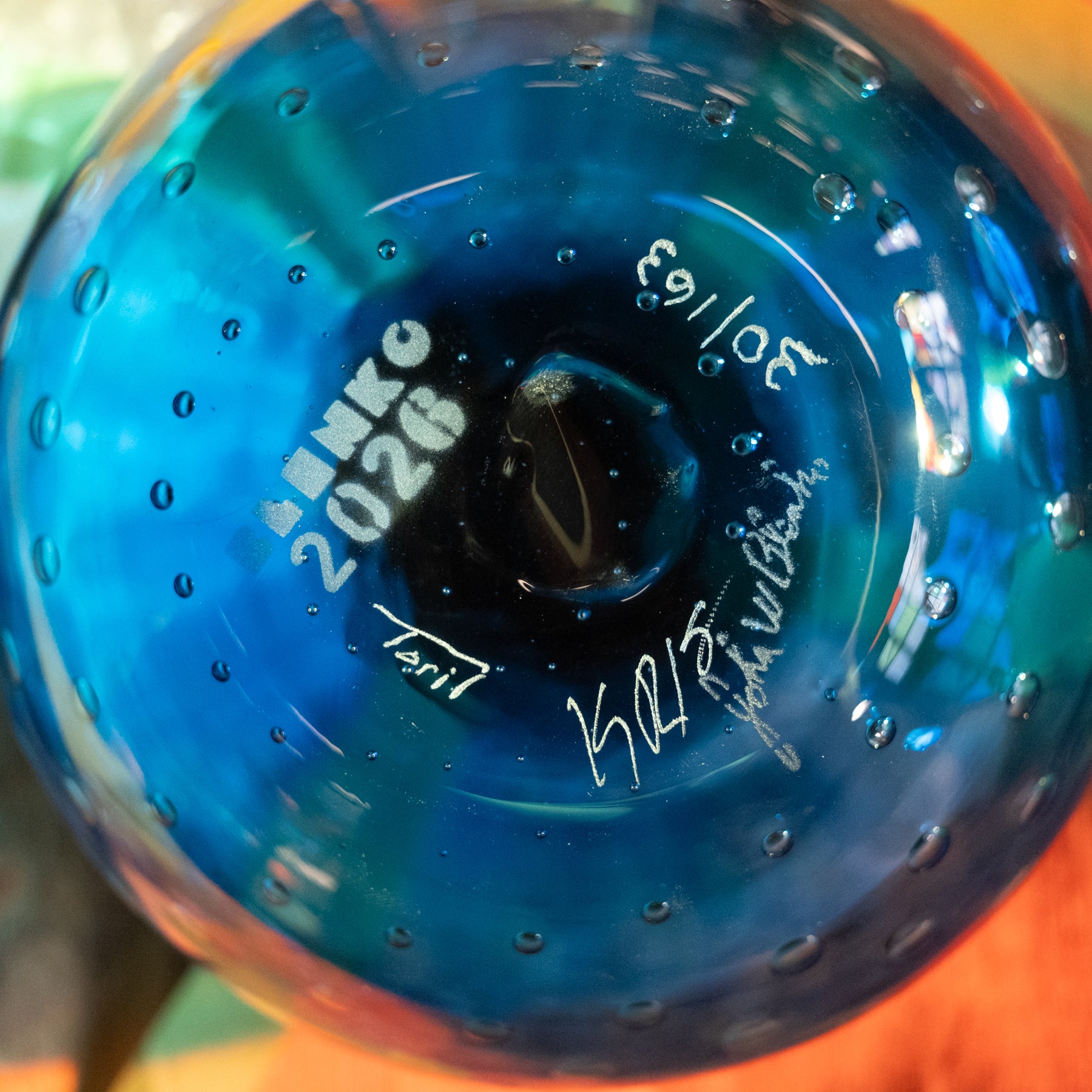 Close-up of a blue transparent glass bottle base with printed white text reading 'AMICO 2026' and signatures, showing water droplets on the glossy surface.