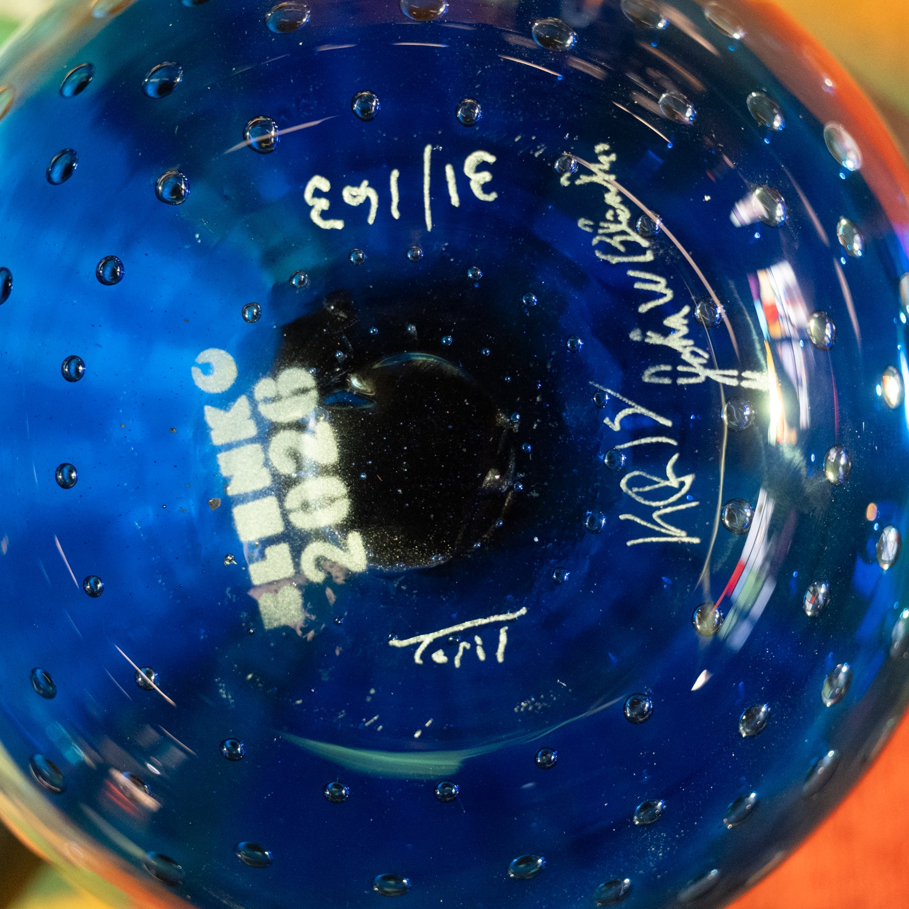 Close-up of the bottom of a blue glass bottle or vase with a bubbled texture, featuring white printed text reading 'SILKMO 2026 3/16/63' and white handwritten signatures.