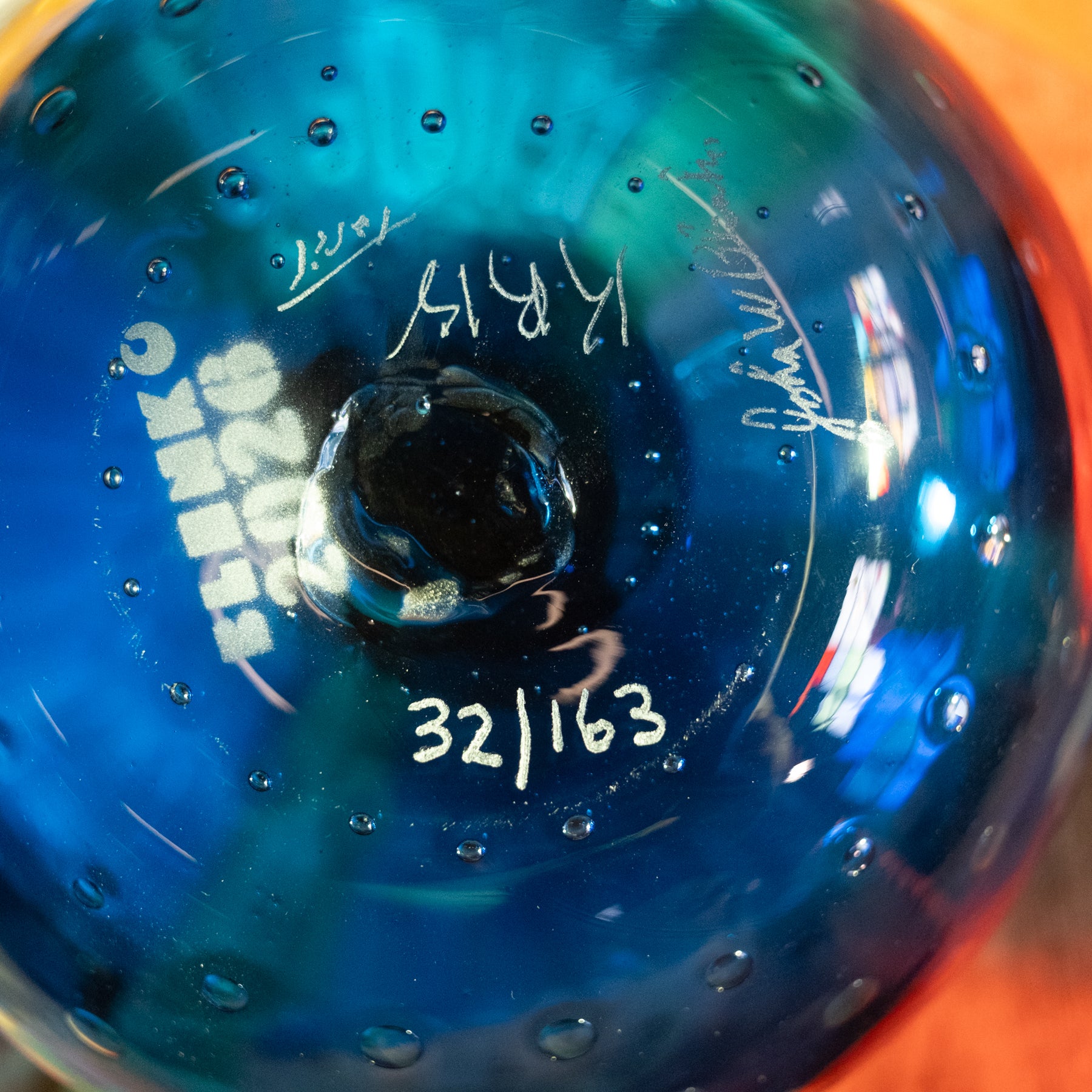 Close-up of the inside of a blue glass bowl or vase with white handwritten markings, including 'KIKO', '32/163', and signatures, showing a glossy textured surface with air bubbles.