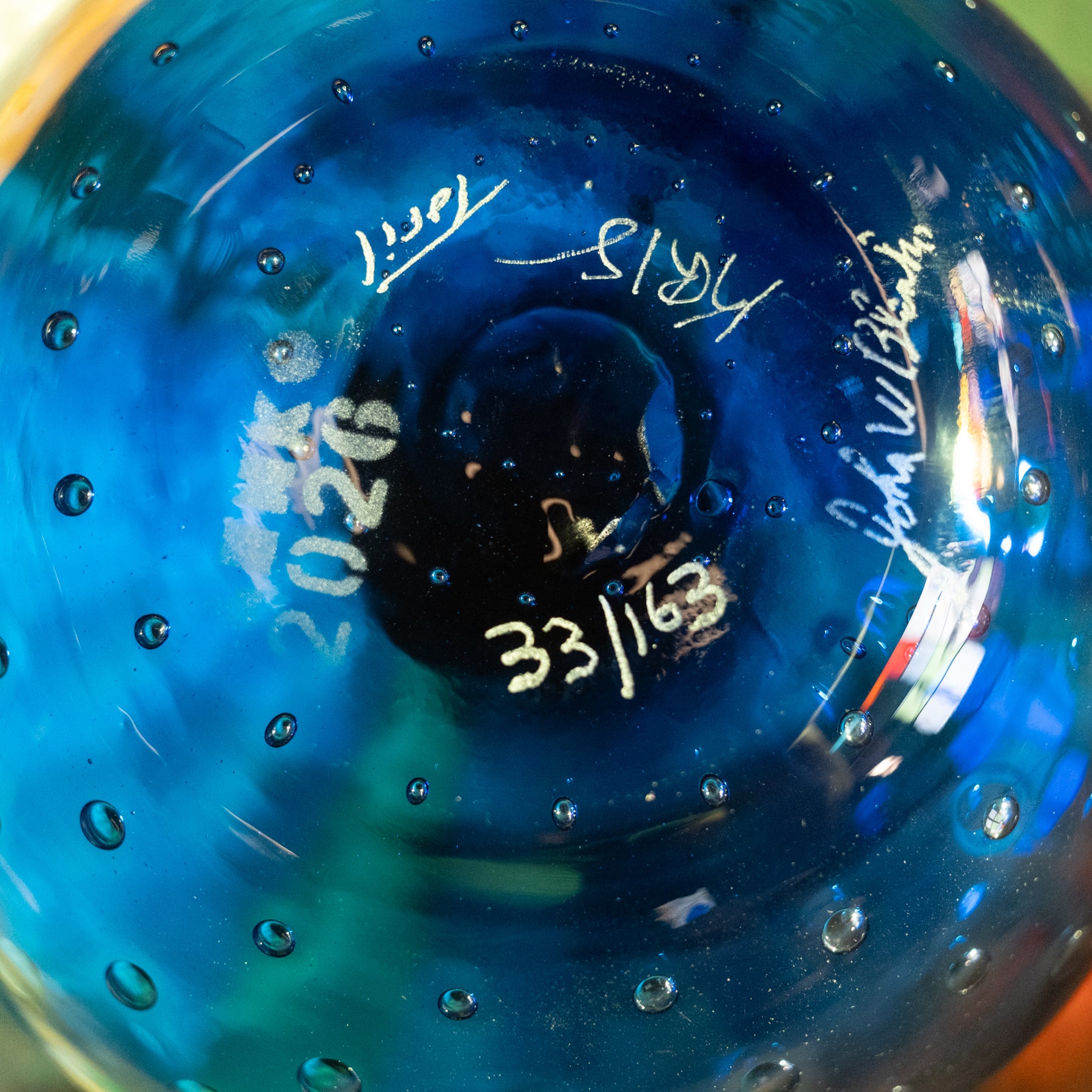 Close-up of the inside of a blue glass bowl or vase with bubble texture, white handwritten dates and signatures, and a dark circular center.