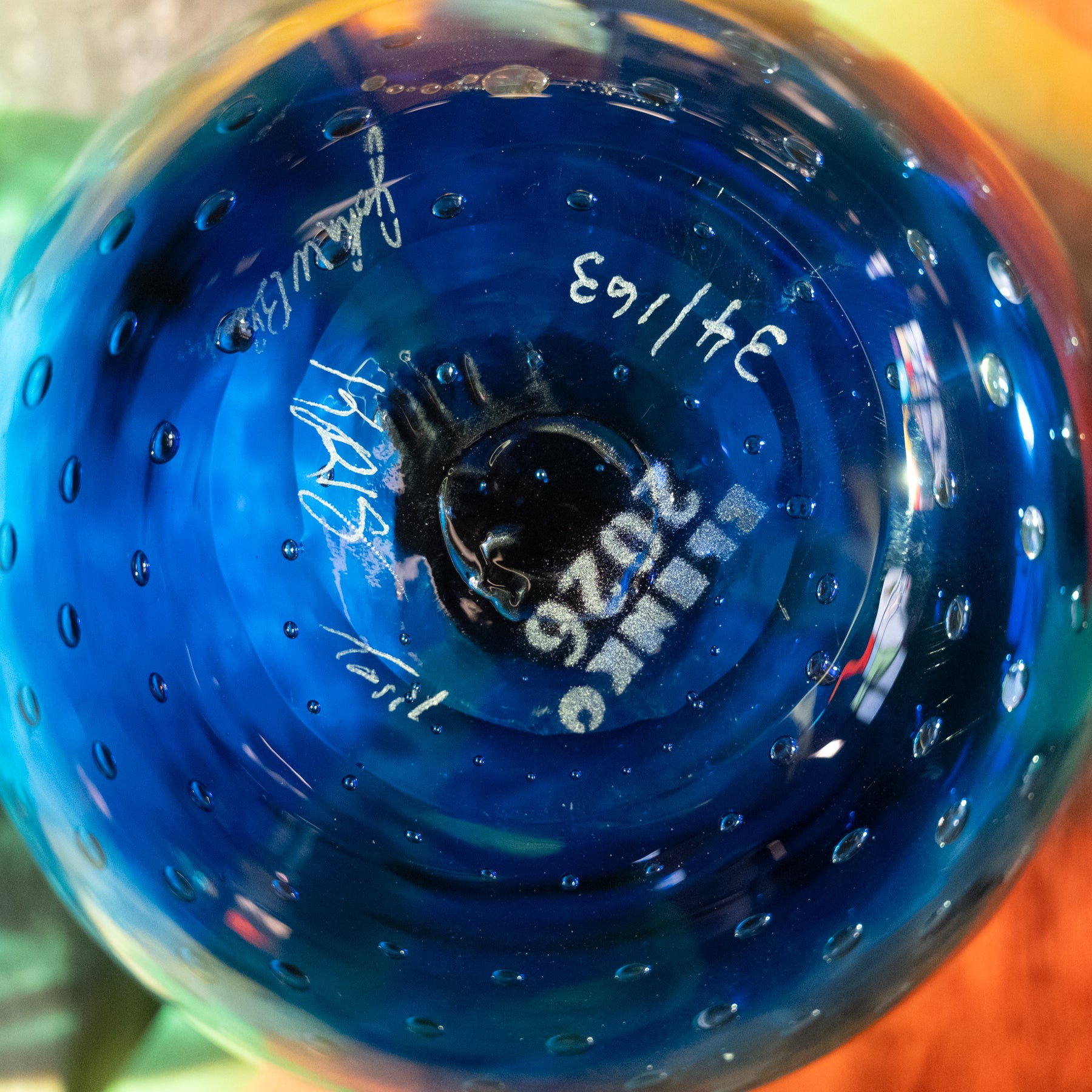 Close-up of the bottom of a transparent blue glass bowl or vase with bubble perforations and white printed markings including 'LIMO' and handwritten numbers.