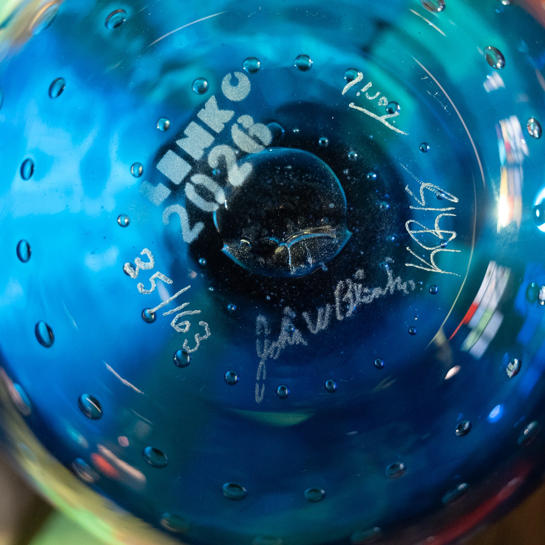 Close-up of the bottom of a blue glass bottle or vase with embossed text reading 'LINKS 2026', small bubble-like perforations around the edge, and white handwritten signatures and dates on the surface.