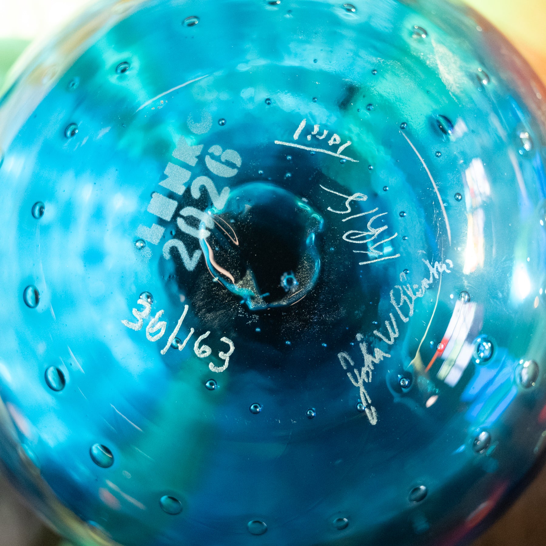 Close-up of a transparent blue glass bottle or vase with embossed text reading 'TENNIS 2026' and white handwritten dates and signatures on the inside surface.