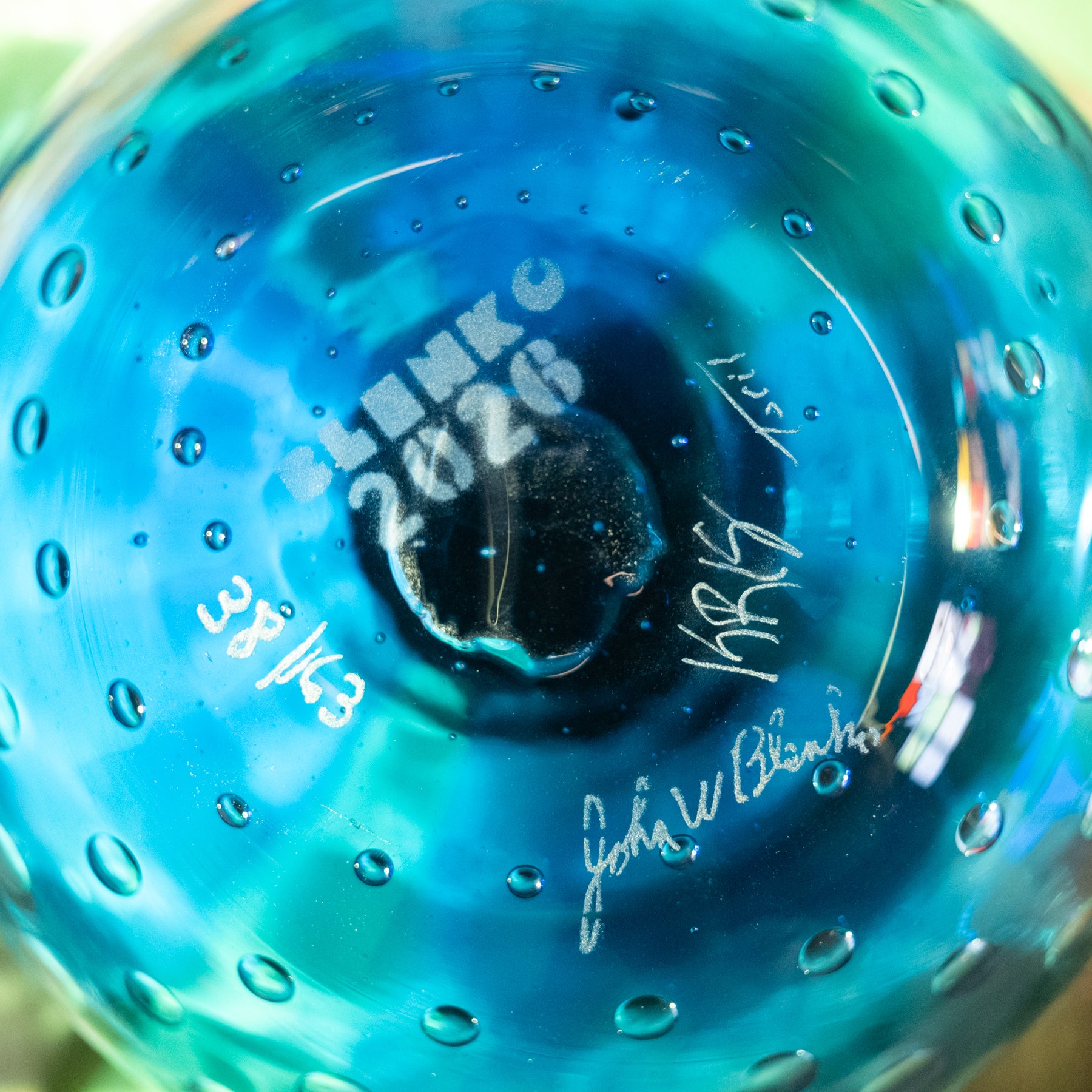 Close-up of a blue and turquoise glass bowl with a textured, bubble-covered surface and white printed text in the center reading 'BLENKO 2026'.