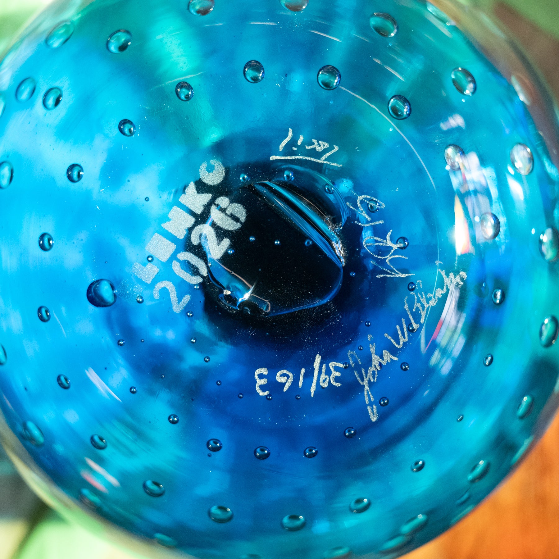 Close-up of a blue glass bottle or vase with a rounded shape, covered in small raised bubble-like dots, showing white printed text and signatures on the bottom.