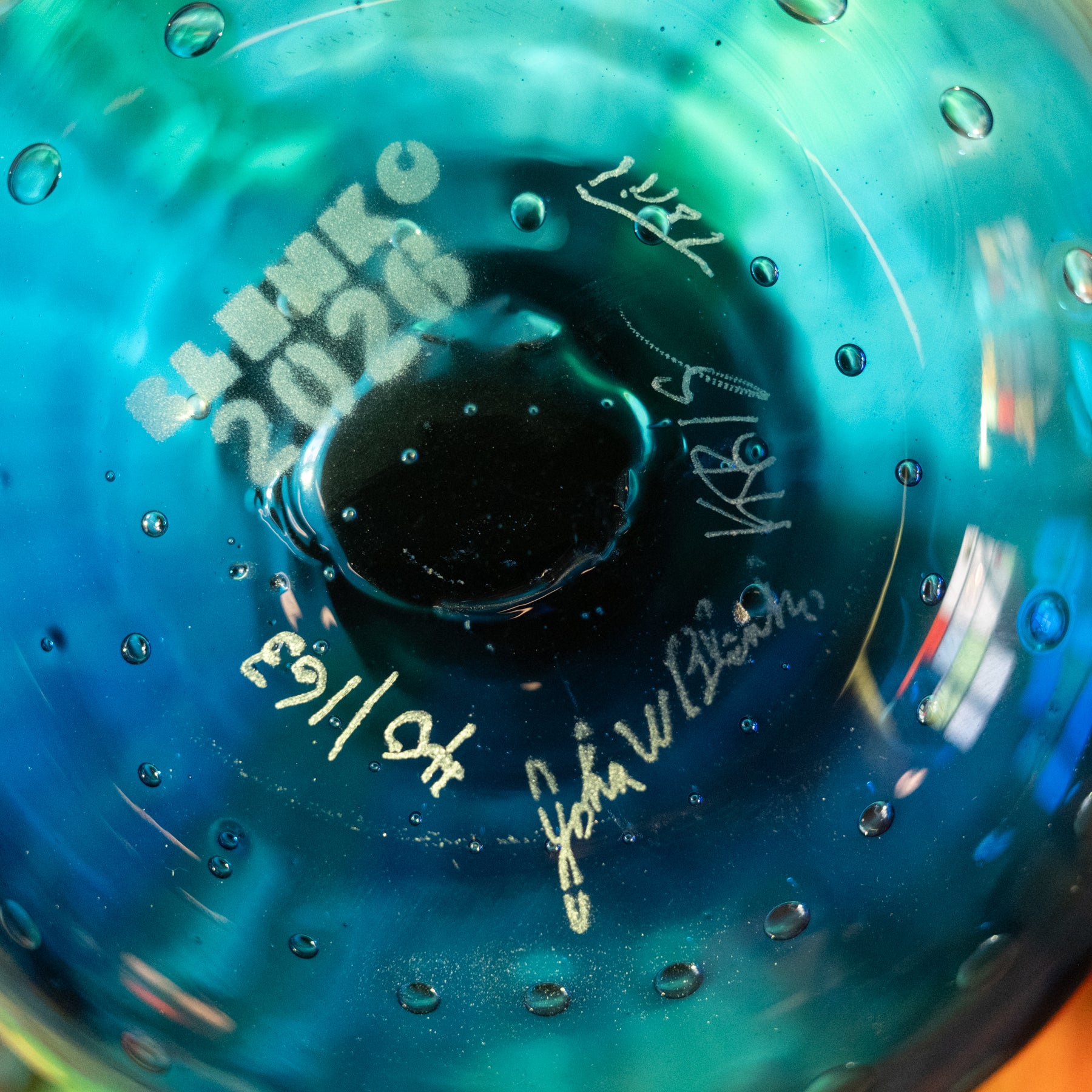 Close-up of a blue and teal glass bowl or vase with embossed text reading 'EL BILBO 2026' and several white handwritten signatures or markings on the inside surface.