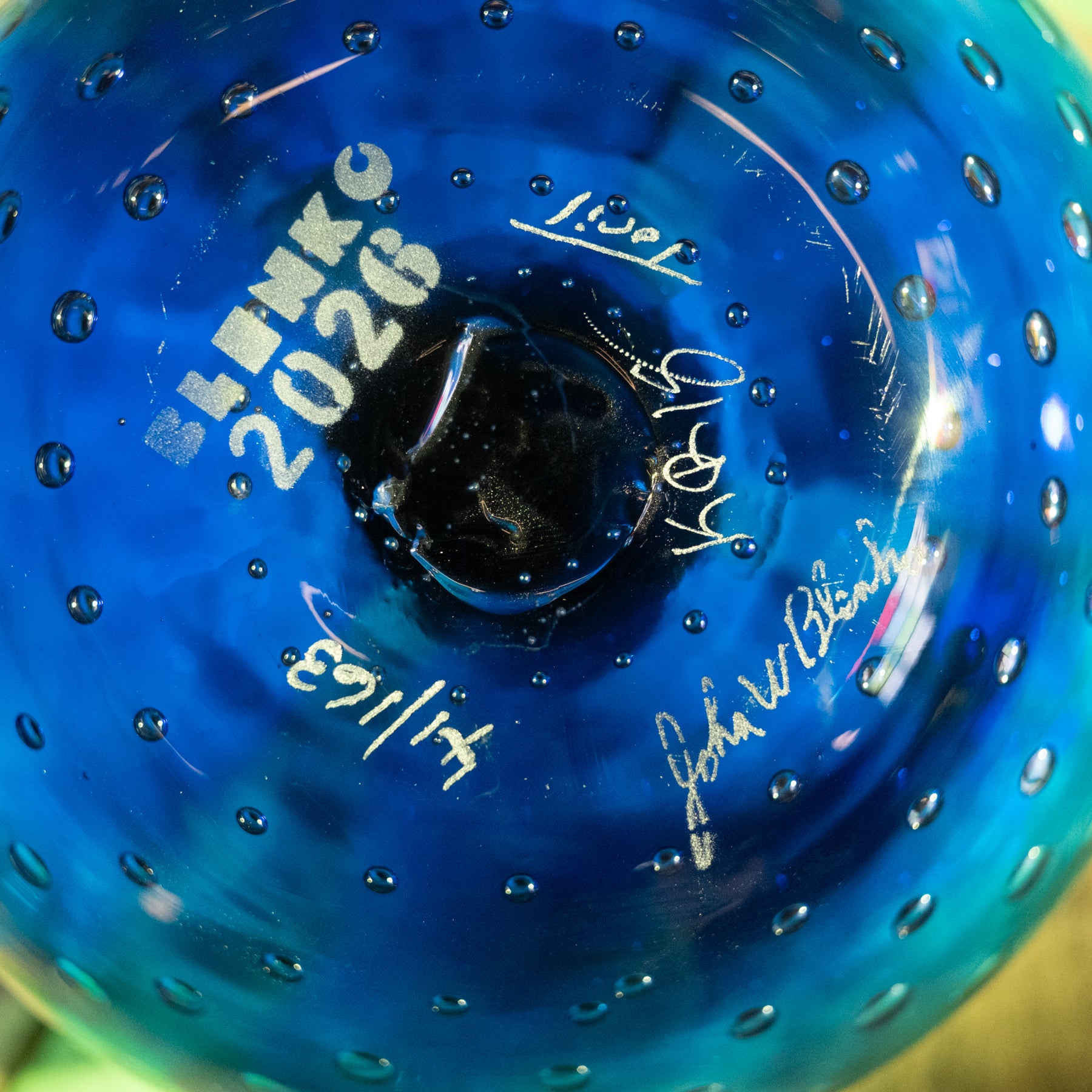 Close-up of a blue glass tumbler with a textured bubble pattern, featuring white printed text reading 'BLENK 2026' and several white signatures.