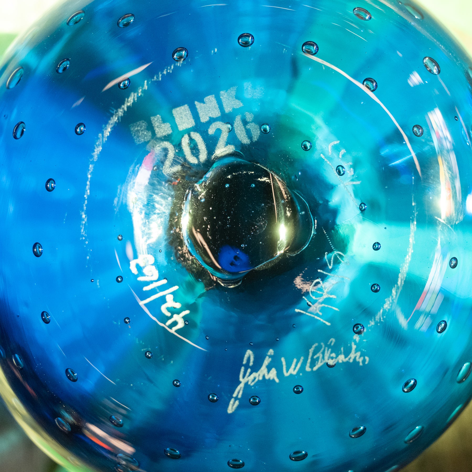 A close-up of a blue glass ornament or decorative piece with a central dark void, featuring white printed text reading 'BLANK 2026' and several white signatures, including 'John W. Blank'.