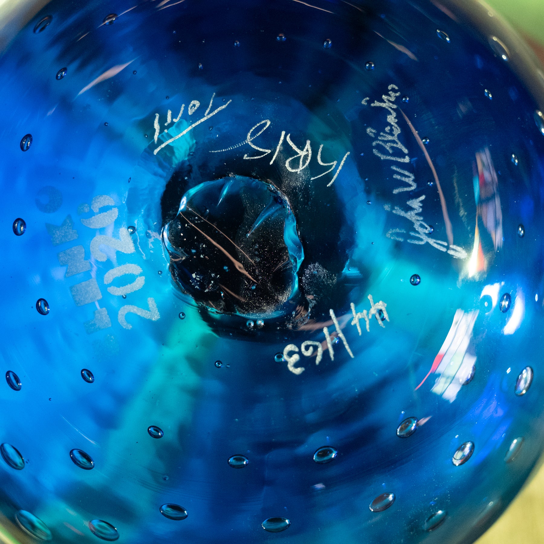 Close-up of a blue glass decorative object with a textured bubble pattern and white handwritten markings and signatures on the inside.