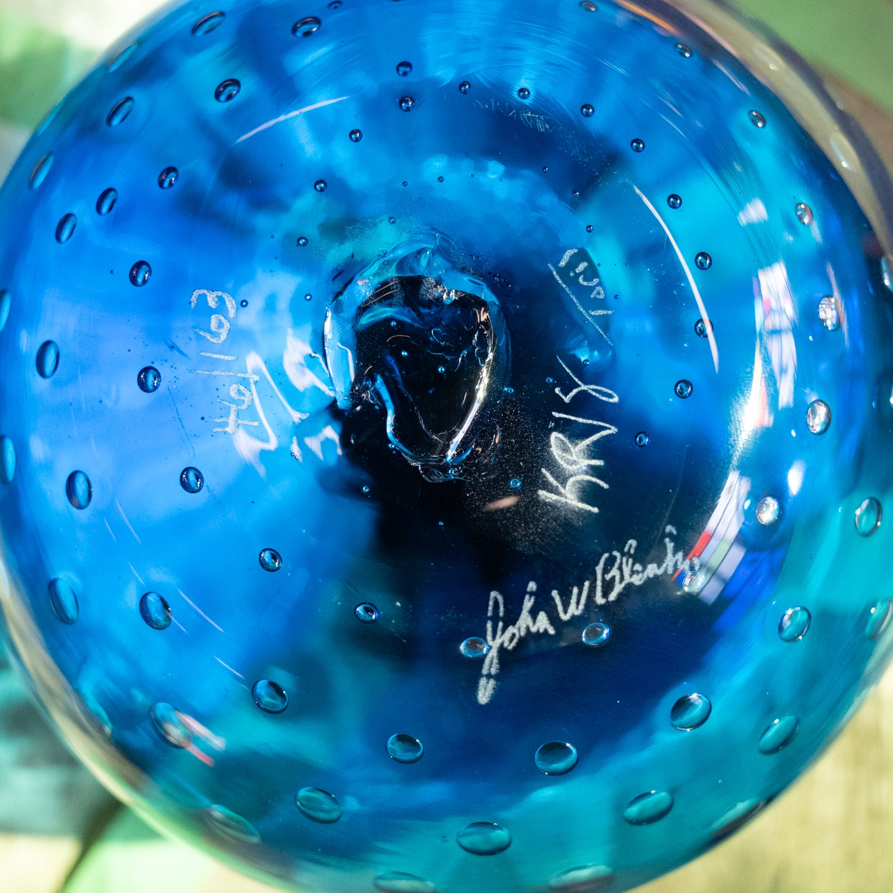 A close-up of a round blue glass bowl with a textured bubble pattern and white artist signatures printed on the side.