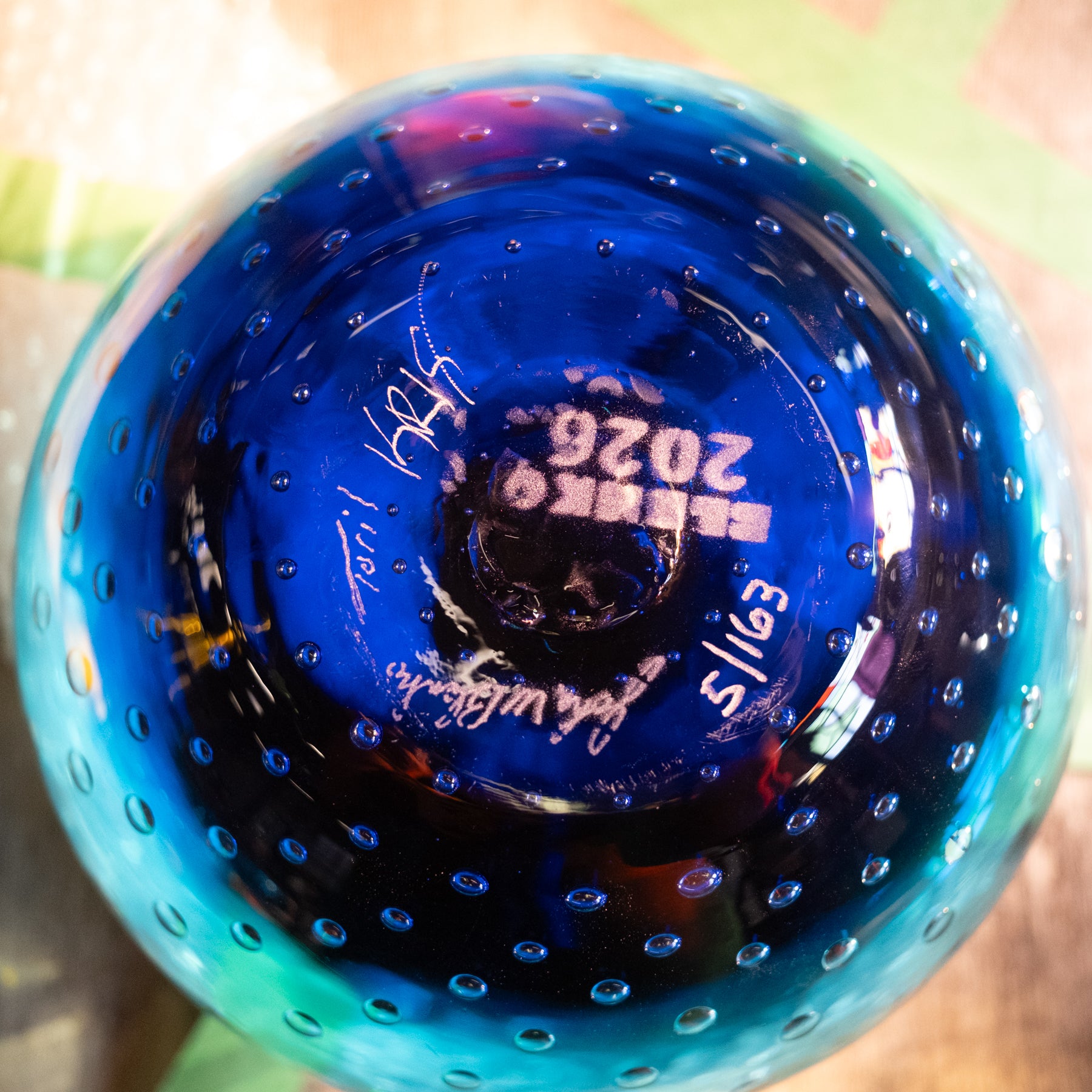 A close-up shot of the bottom of a hand-blown glass bowl, showcasing its deep cobalt blue color with lighter turquoise-green edges, a textured bubble pattern, and white hand-written signatures.