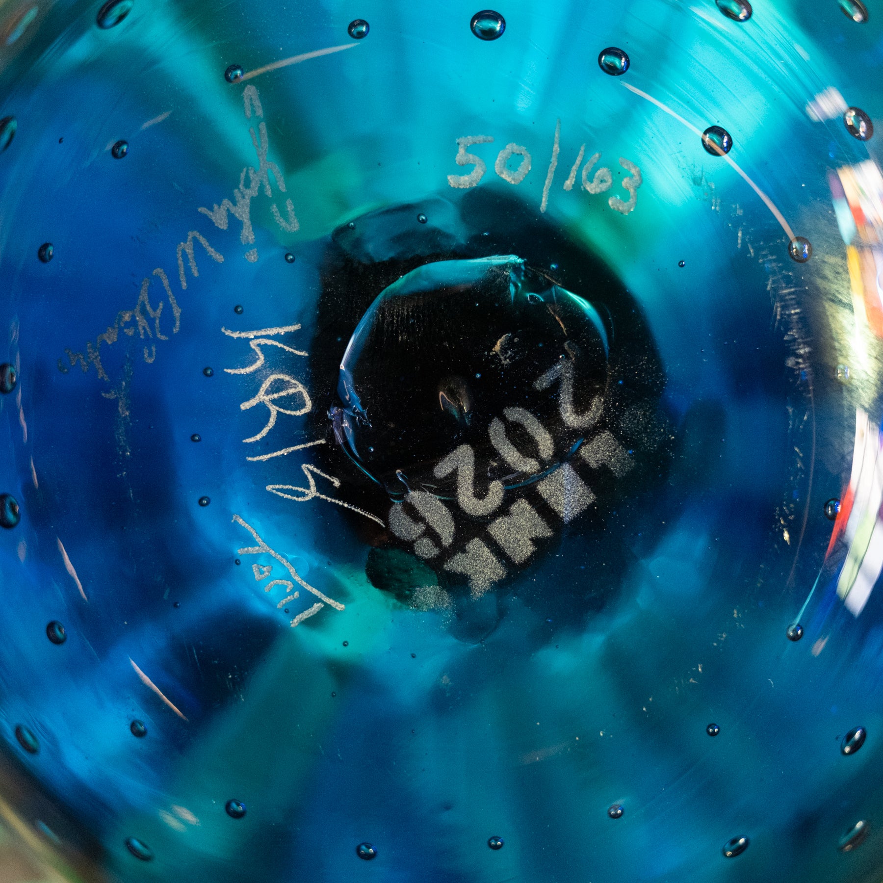 A close-up of a blue glass bottle or vase viewed from inside, showing a dark circular base with white printed text reading 'LINK 2026' and handwritten markings including '50/1103' and signatures.