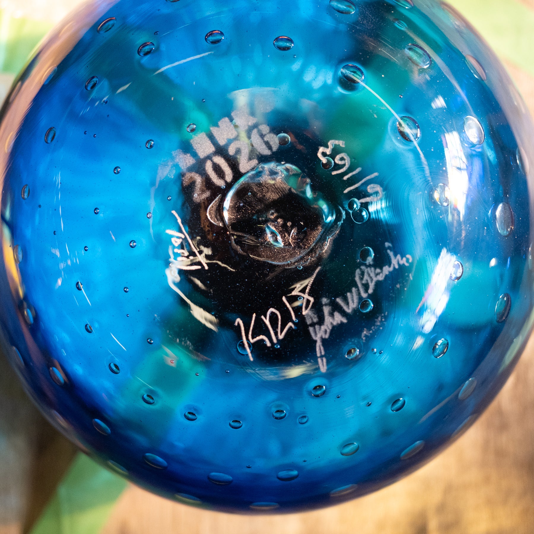 A close-up of a blue glass bottle or vase with a perforated bubble pattern, showing white handwritten markings and signatures on the bottom.