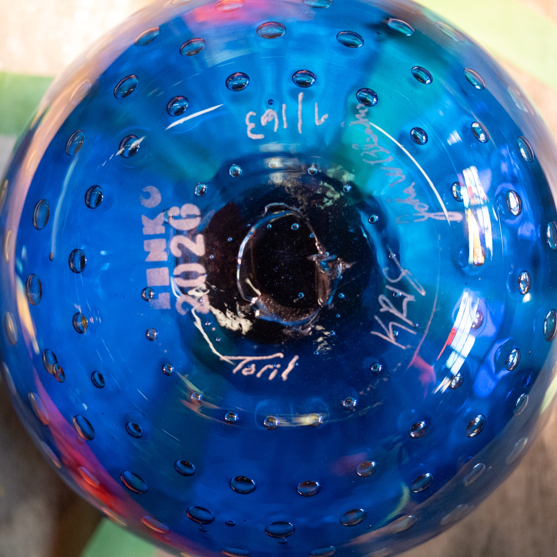Close-up of the bottom of a transparent blue glass bottle with a perforated bubble pattern, showing white printed text and signatures.