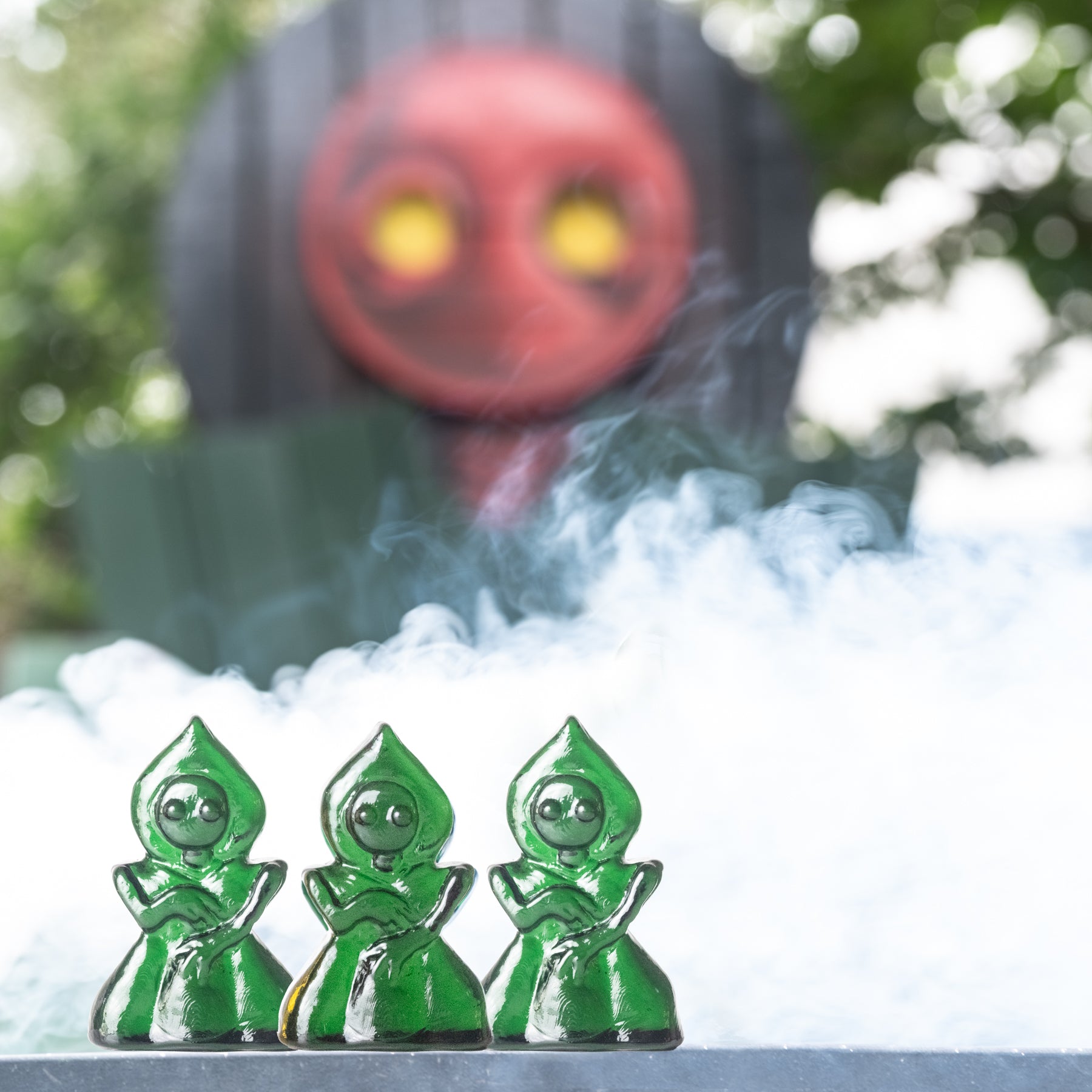 Three green hooded figures with a red face in the background, surrounded by smoke.