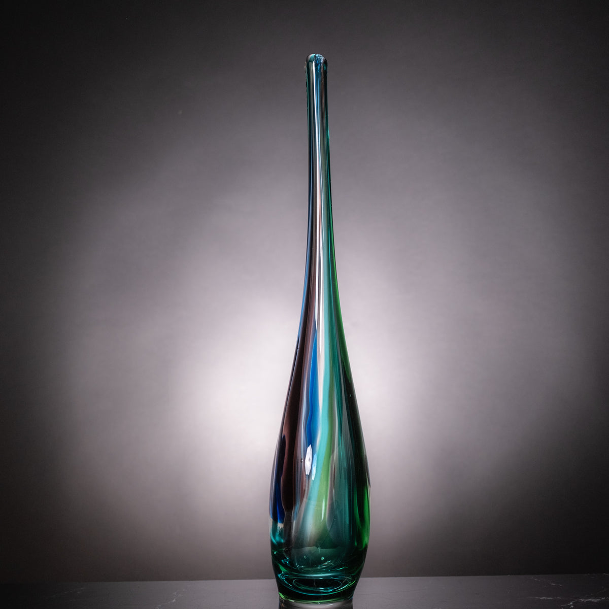 A tall, slender glass vase with a teardrop shape, featuring iridescent swirls of green, blue, and purple, set against a dark gray gradient background.