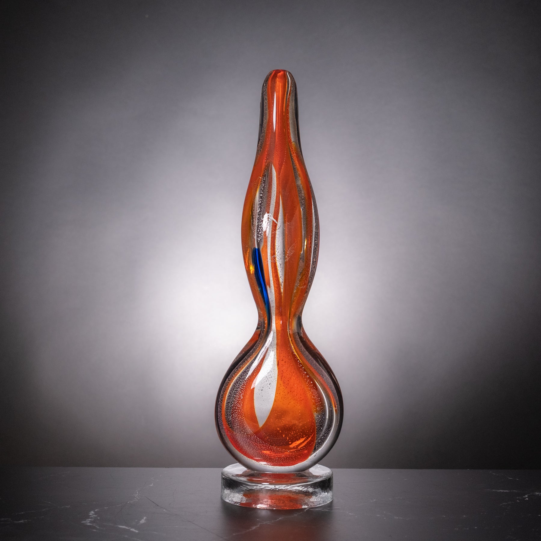 A sculptural glass sculpture with a wavy, flame-like silhouette, featuring vibrant orange and red glass swirls with a small blue accent, displayed on a clear glass base against a dark gray gradient background.