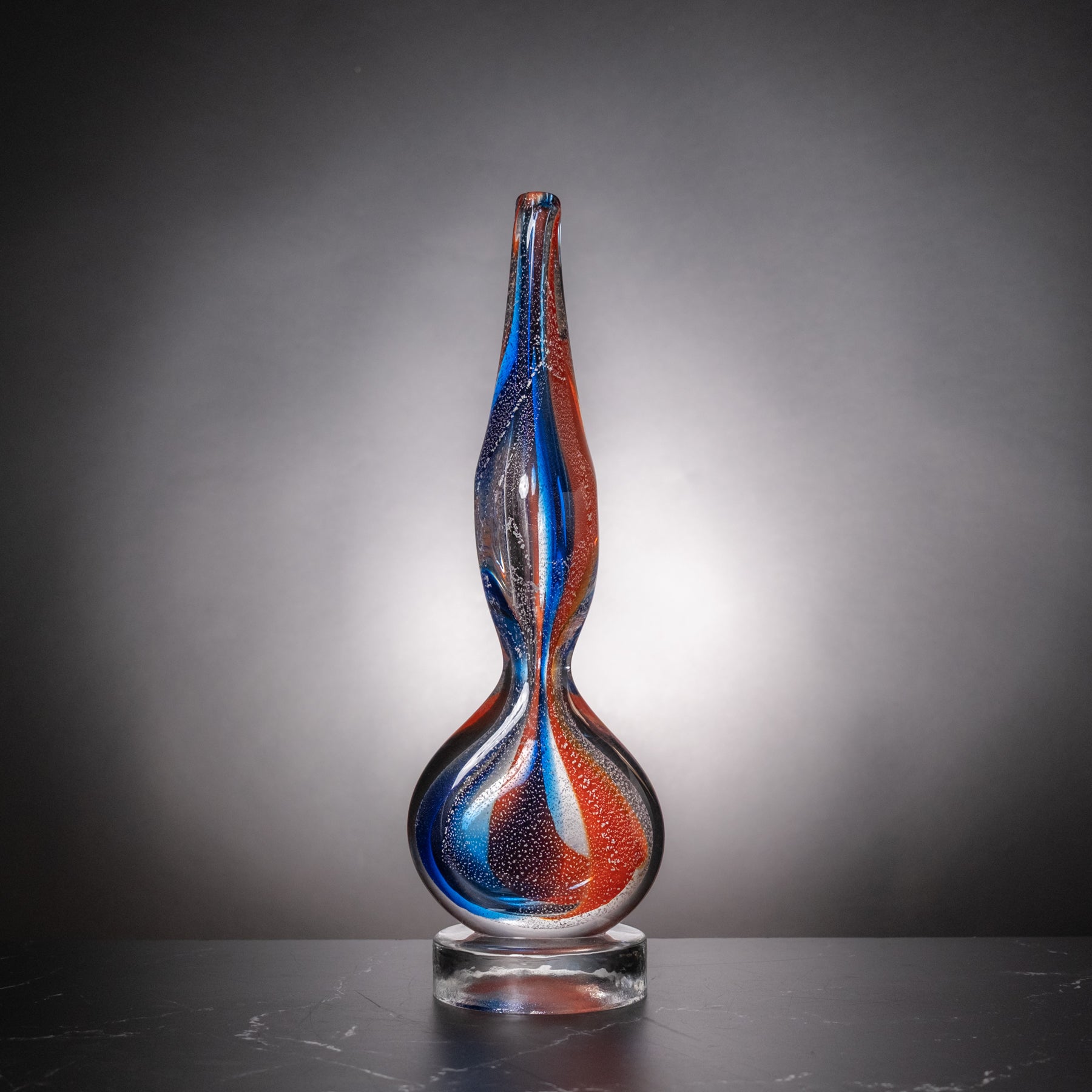 A tall, sculptural glass vase with a bulbous base and a narrow neck, featuring swirls of blue, red, and clear glass, displayed on a clear glass stand against a dark gray gradient background.