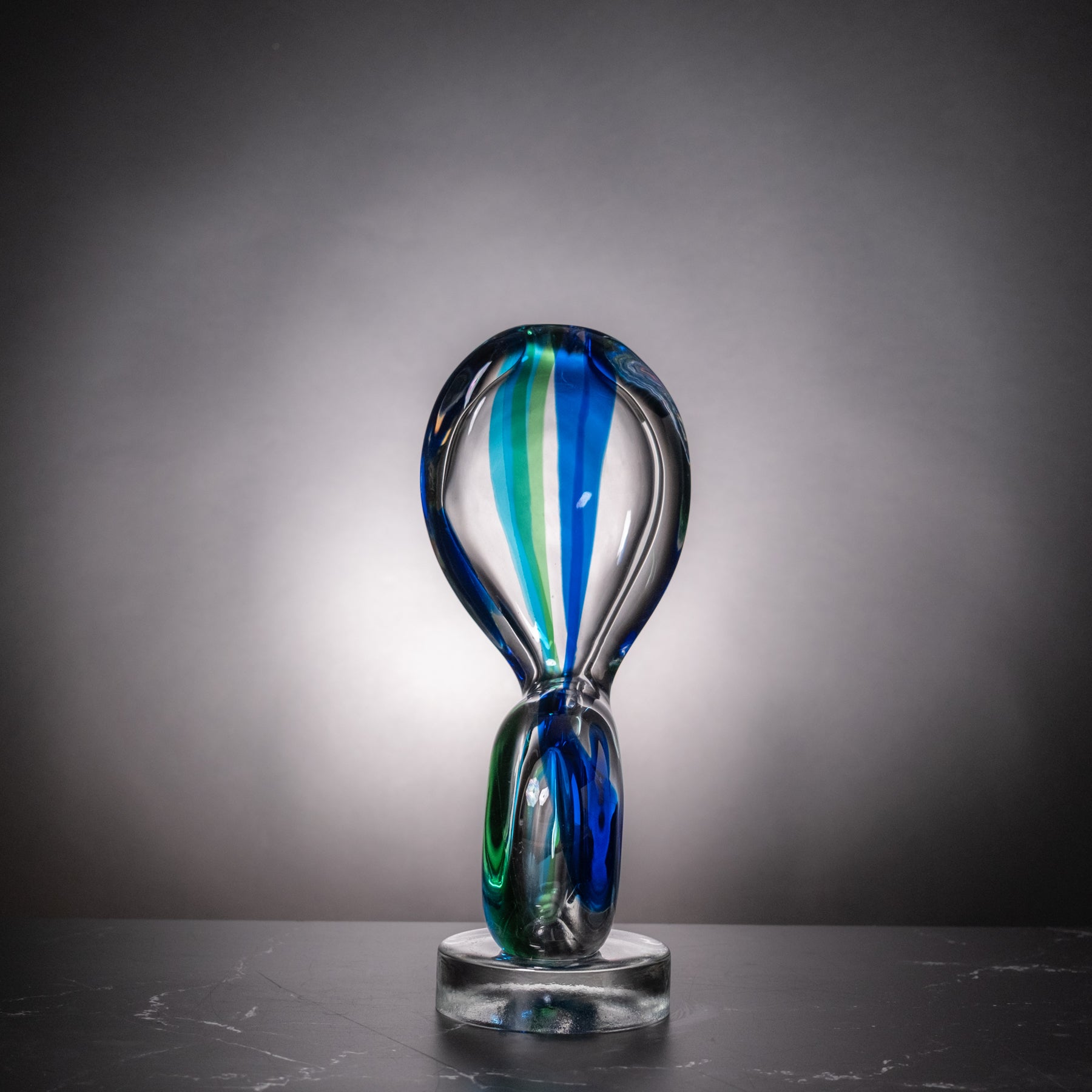 An abstract curved glass sculpture on a clear round base, featuring swirls of blue, green, and clear glass, set against a dark gray gradient background.