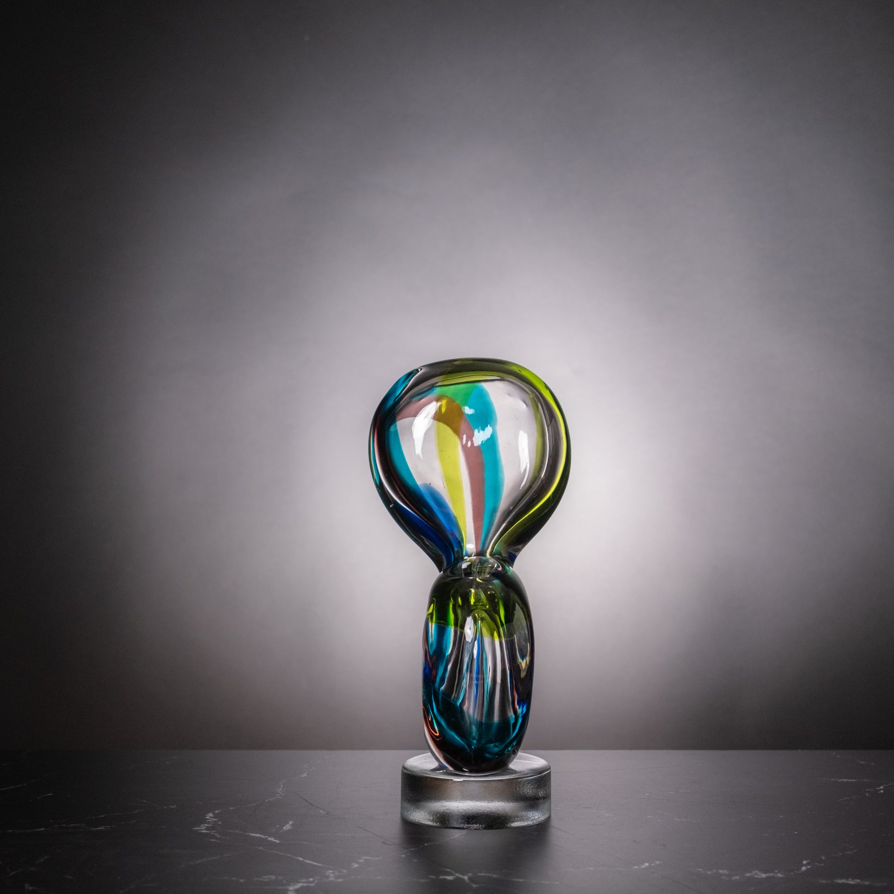 A modern abstract glass sculpture with a looped top and a clear cylindrical base, featuring swirls of blue, green, yellow, and white glass, set against a dark gray gradient background.