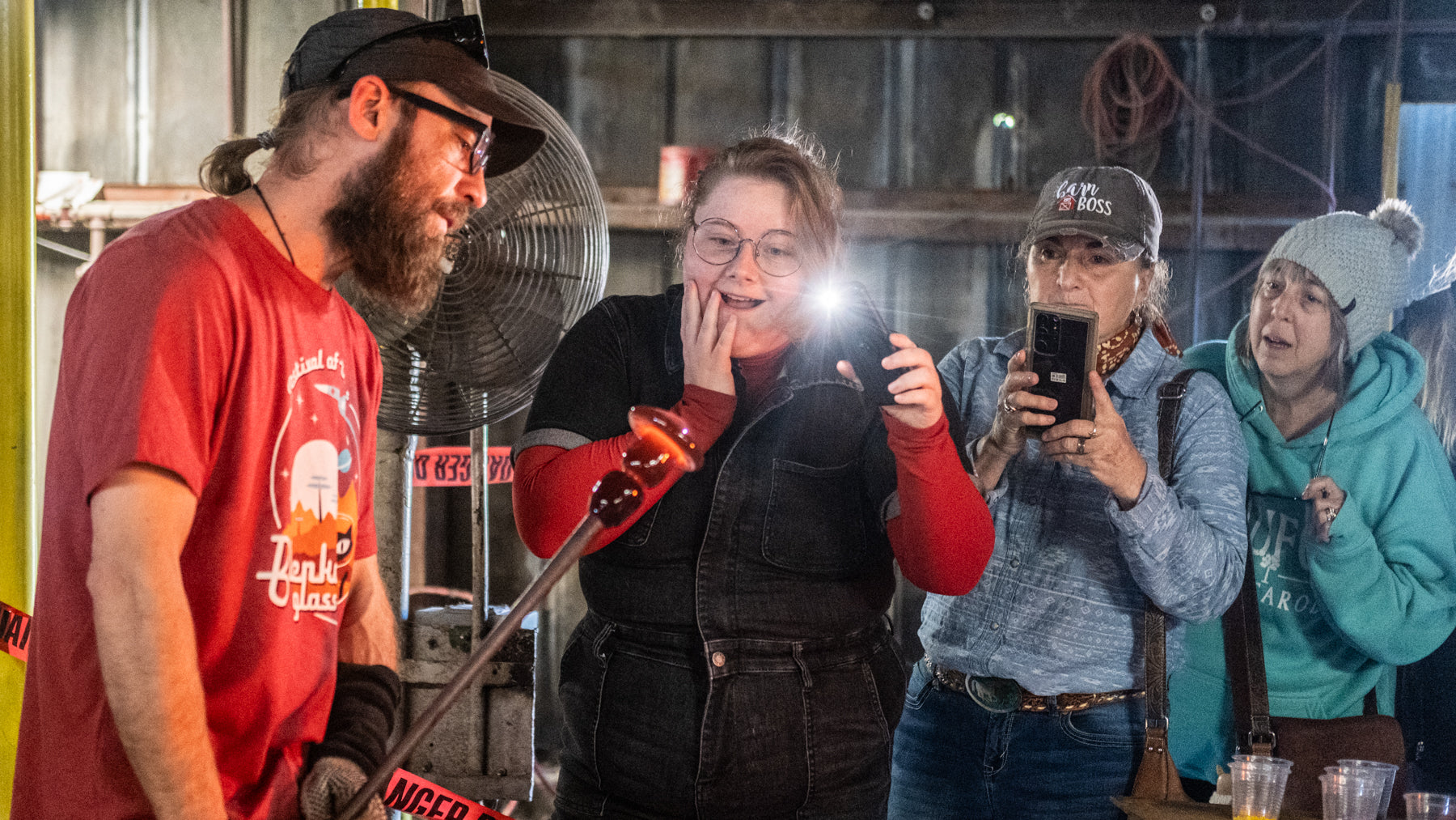 Group of people in a rustic indoor setting, possibly a barn or workshop, with one person holding a phone.