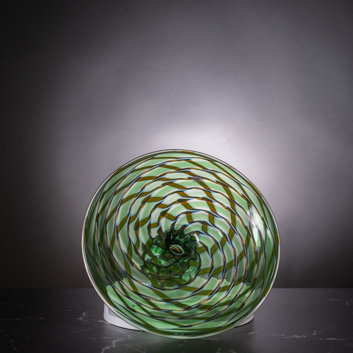 A hand-blown green glass bowl with a spiral pattern of dark brown and blue lines, displayed on a white circular stand against a dark gray gradient background.