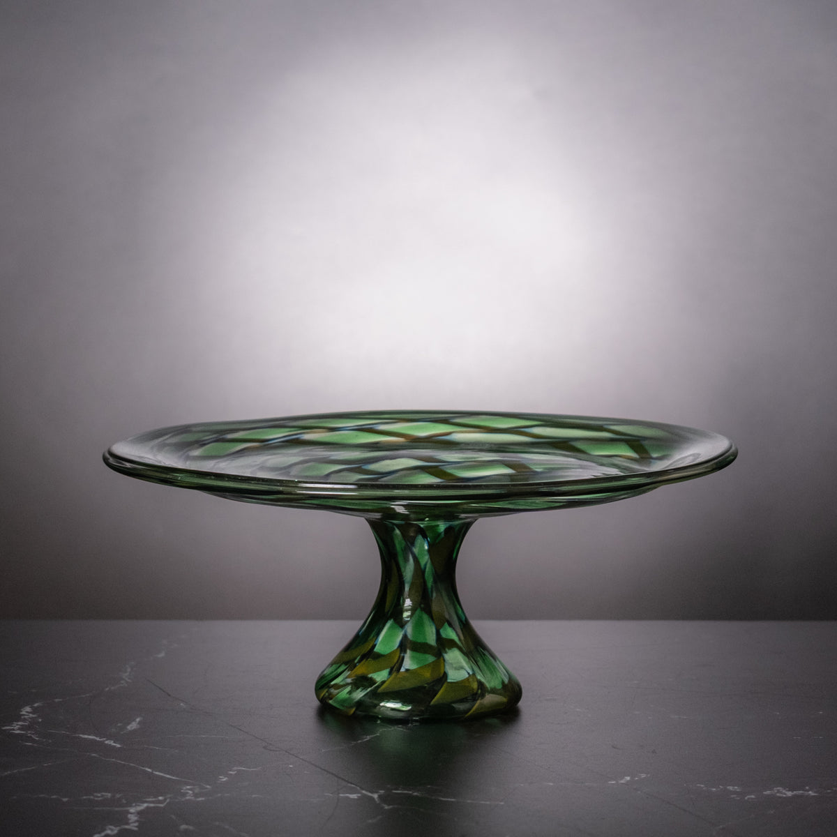 A green glass pedestal plate with a faceted stem and a patterned rim, displayed on a dark marble surface against a gray gradient background.